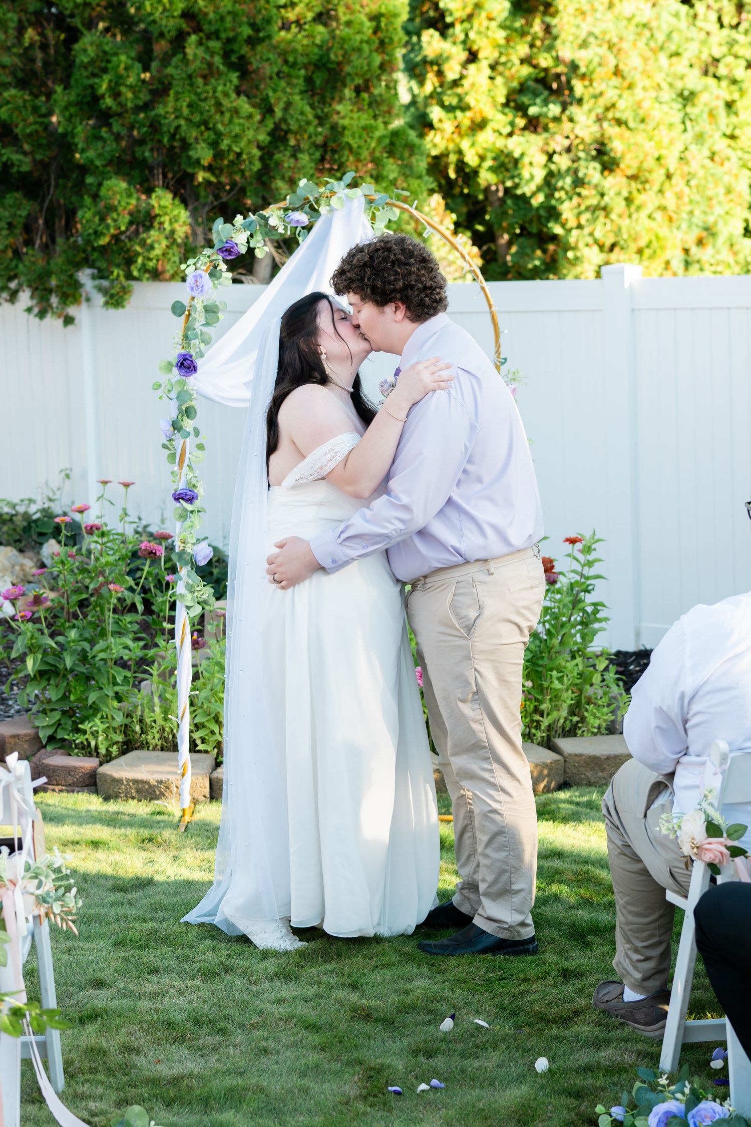 Bride and groom share first kiss at outdoor backyard wedding ceremony under floral hoop arch with white fence backdrop.