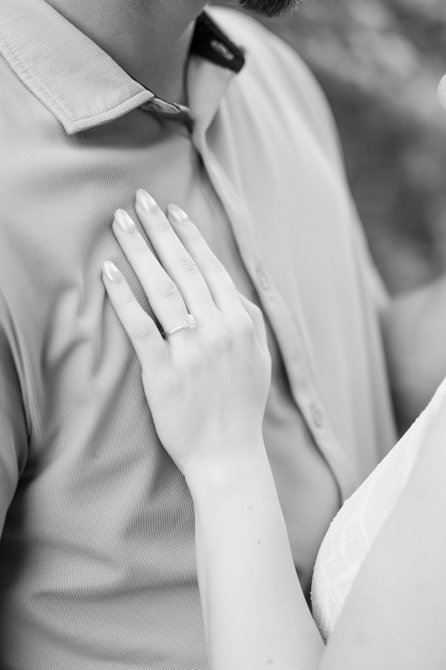 Close-up black and white photo of a woman's hand with engagement ring resting on man's chest.