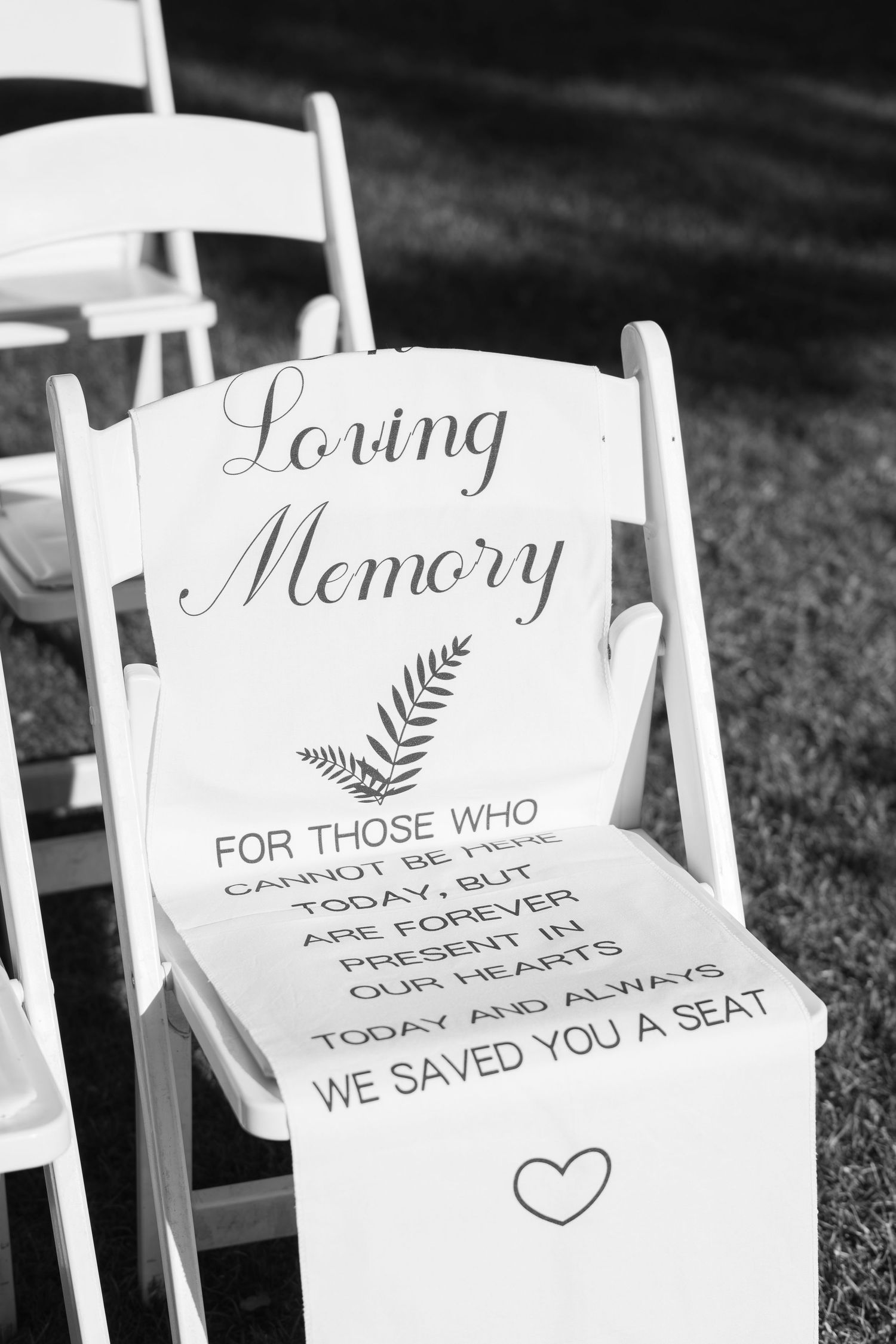 Black and white wedding memorial chair sign reading Loving Memory for absent loved ones at outdoor ceremony.