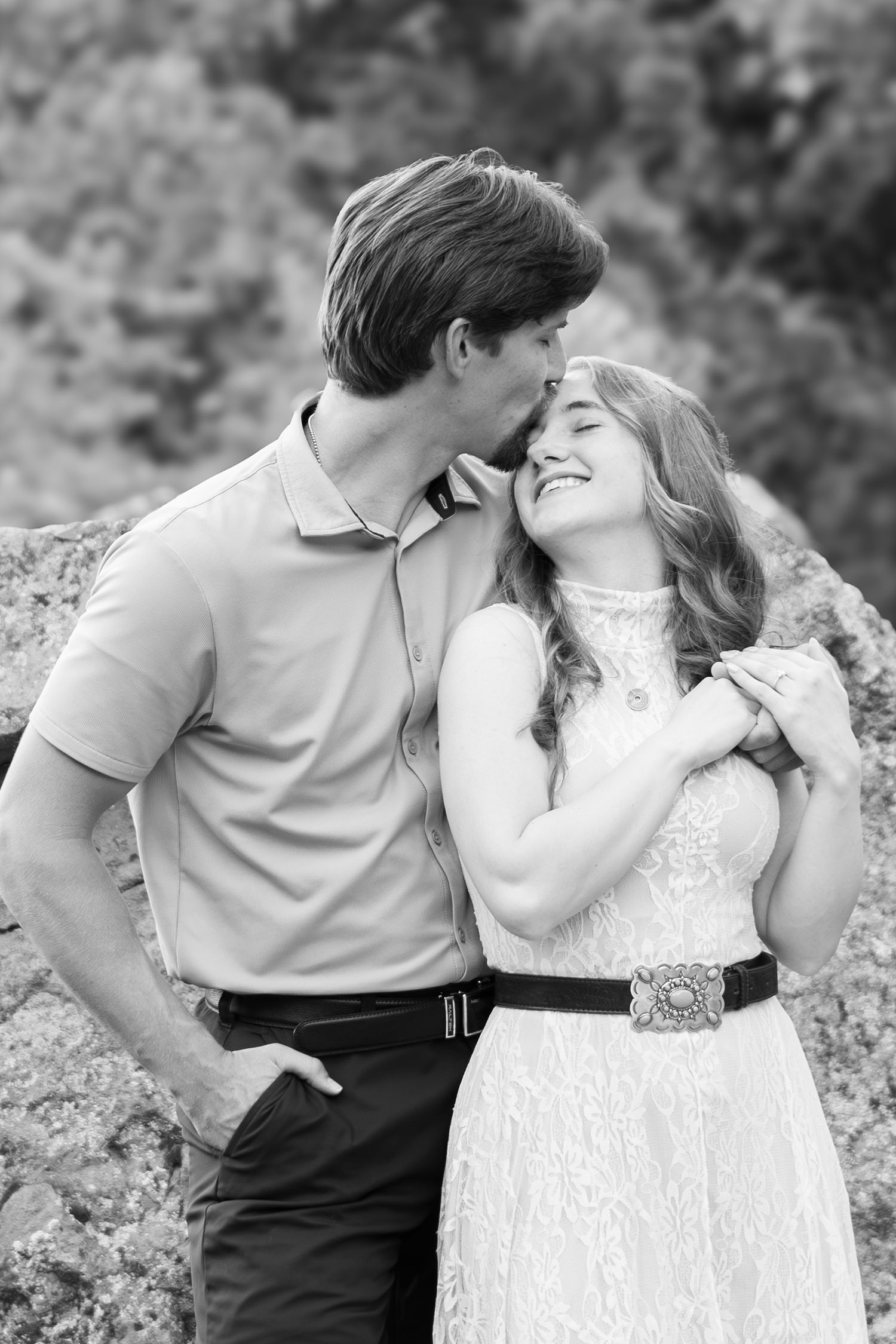 Black and white photo of a couple embracing outdoors, man kissing woman's temple as she smiles warmly.