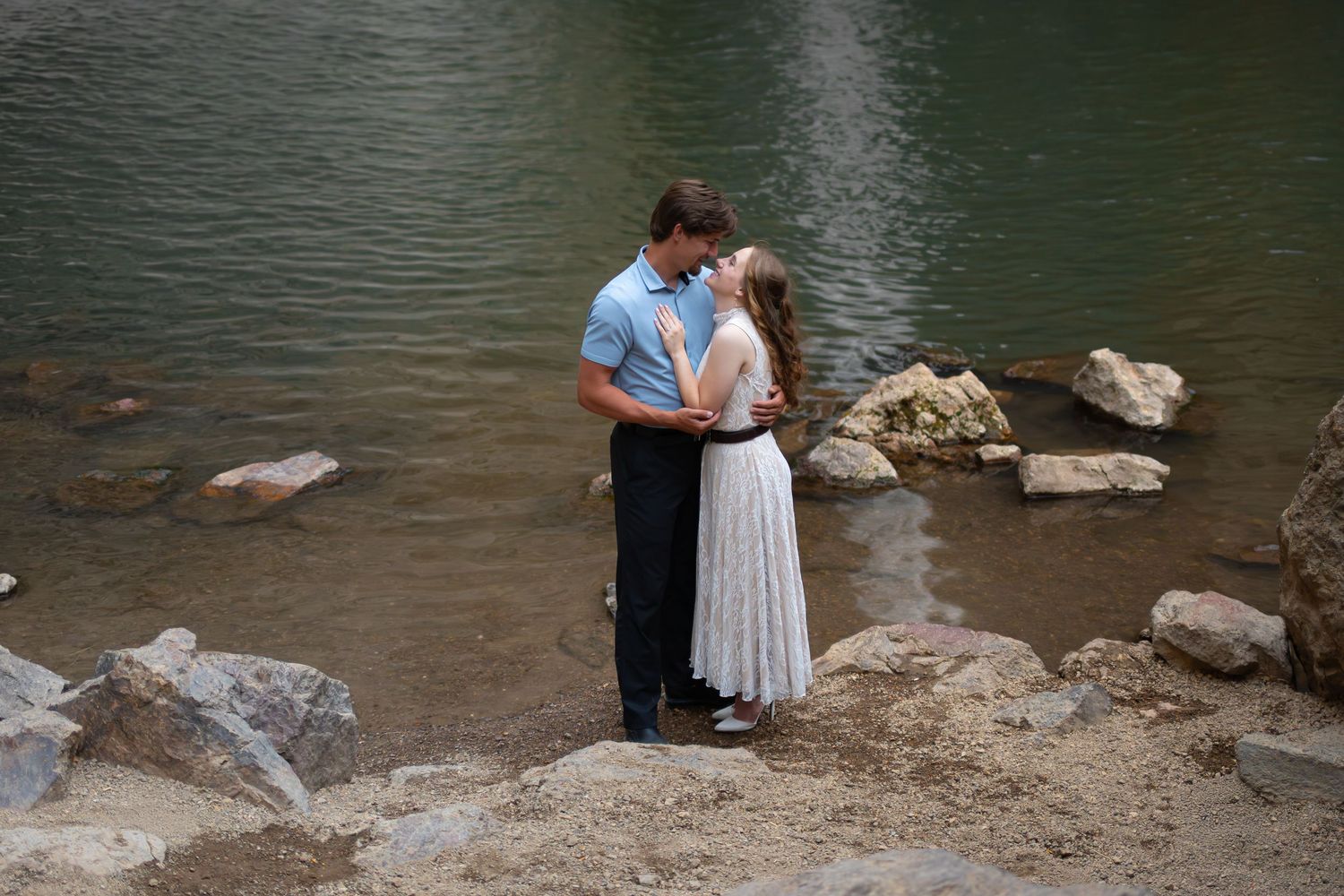 Couple sharing a romantic kiss by a serene rocky lakeside, man in blue shirt and woman in white dress.