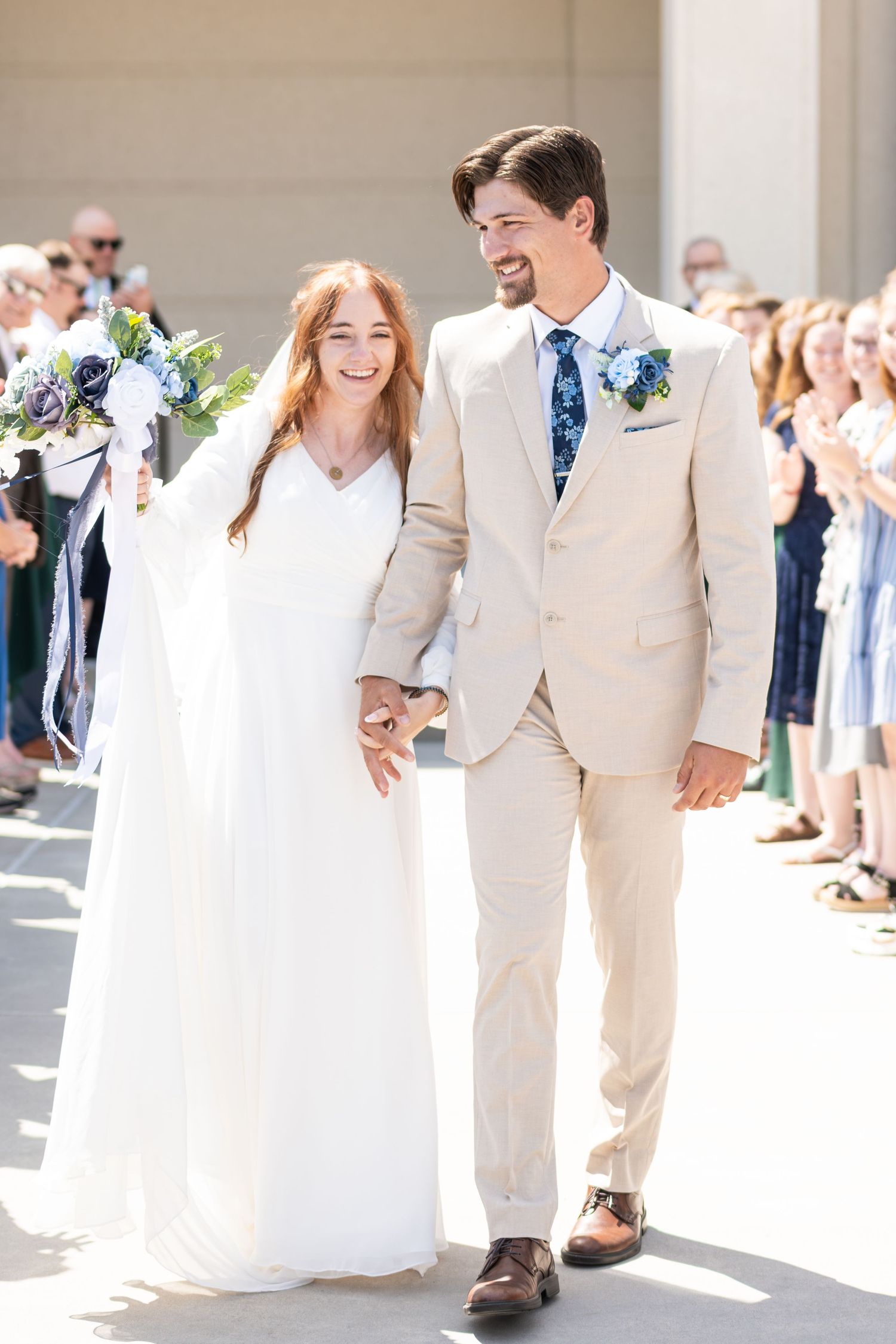 Smiling bride in white gown and groom in tan suit walk hand-in-hand after outdoor wedding ceremony.