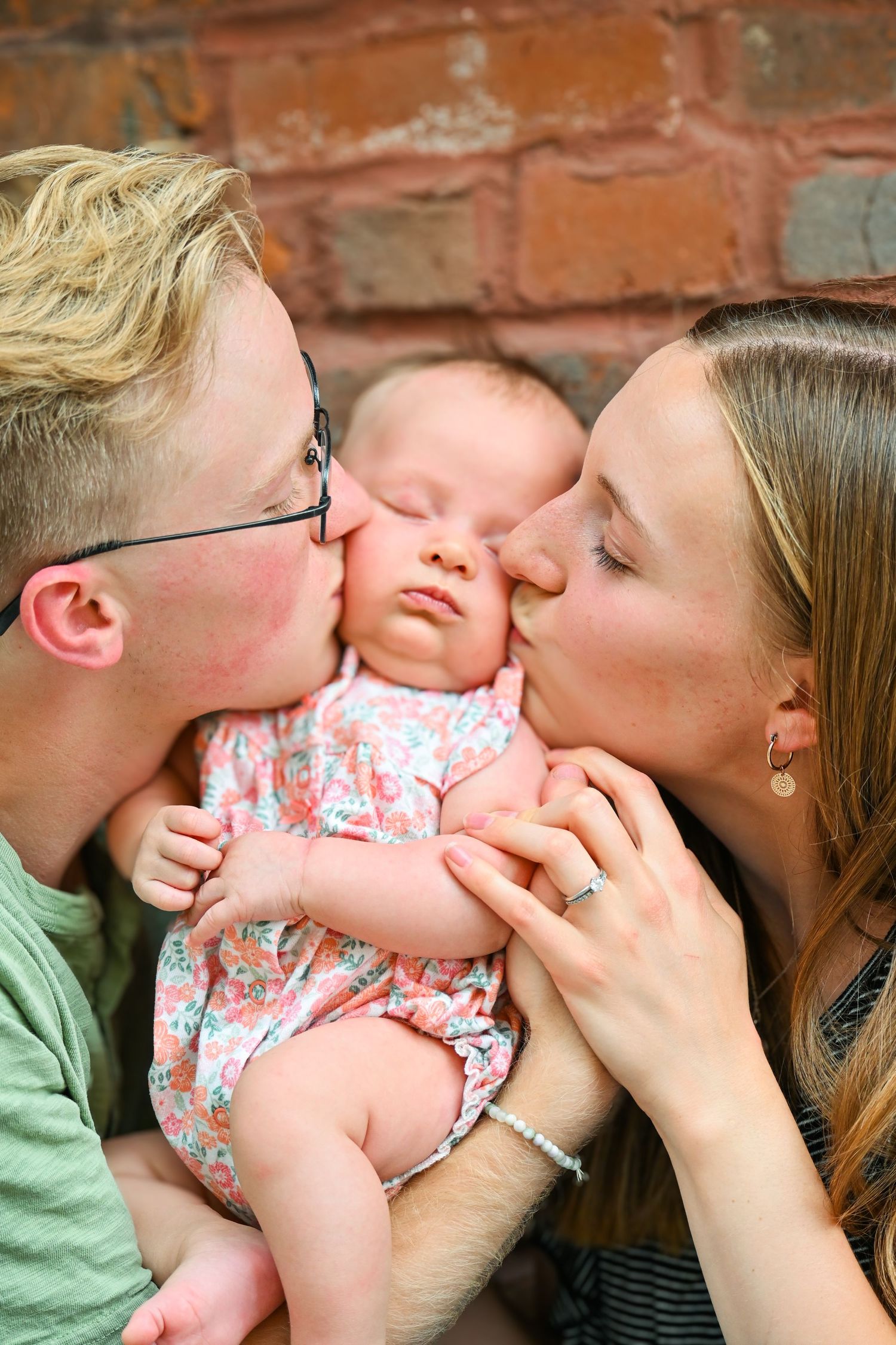 A baby in a floral outfit receives loving kisses from parents against a brick wall background.