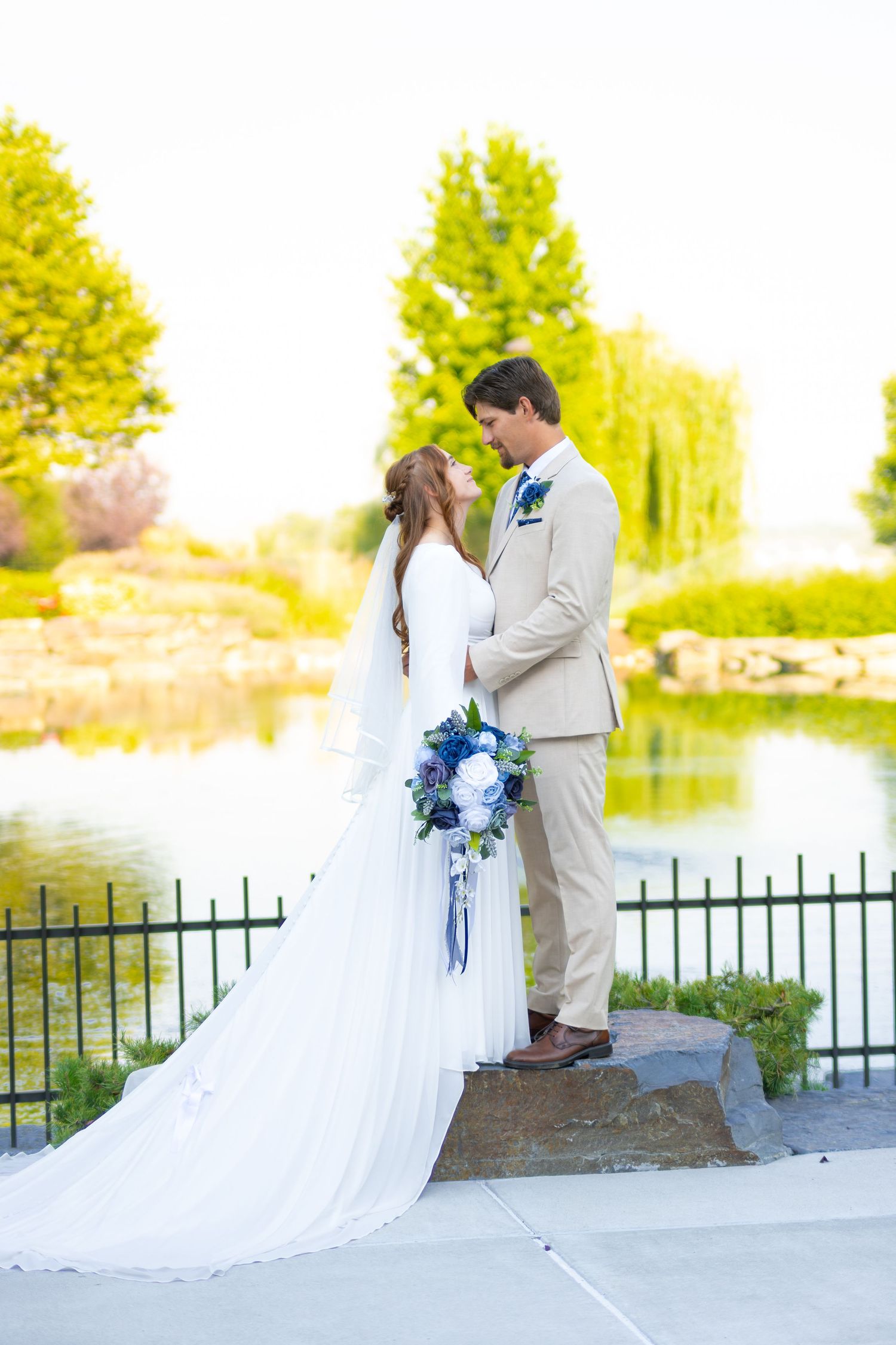 Bride and groom stand facing each other by a pond, she holds blue bouquet, he wears tan suit, lush greenery background.