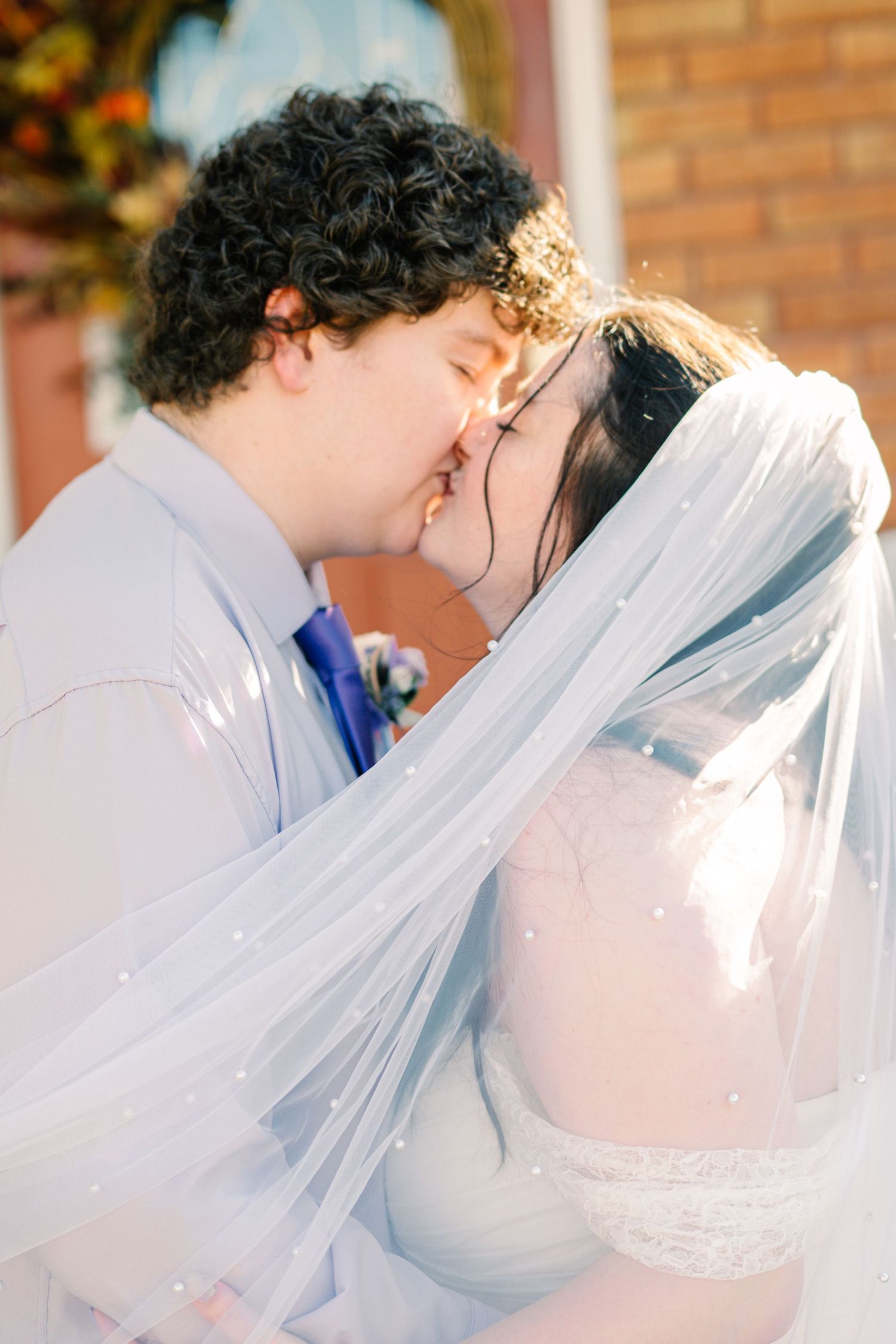 A bride and groom share their first kiss, with the bride wearing a pearl-adorned veil, outdoors near a brick building.
