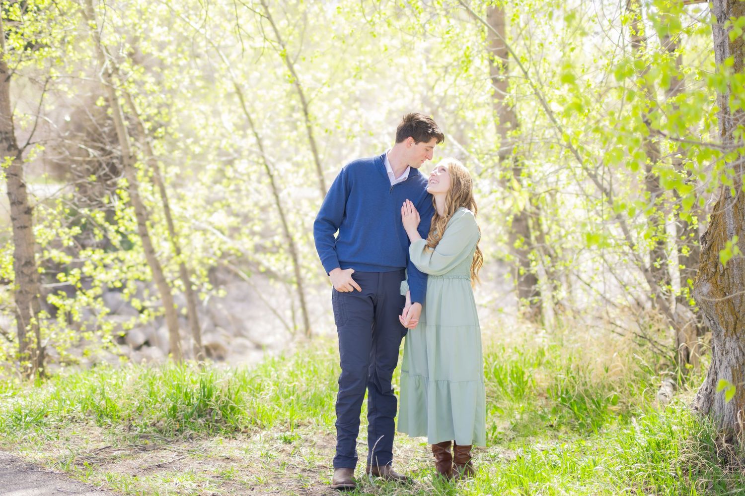 Couple sharing a romantic moment on a sunlit forest path surrounded by lush green spring trees.