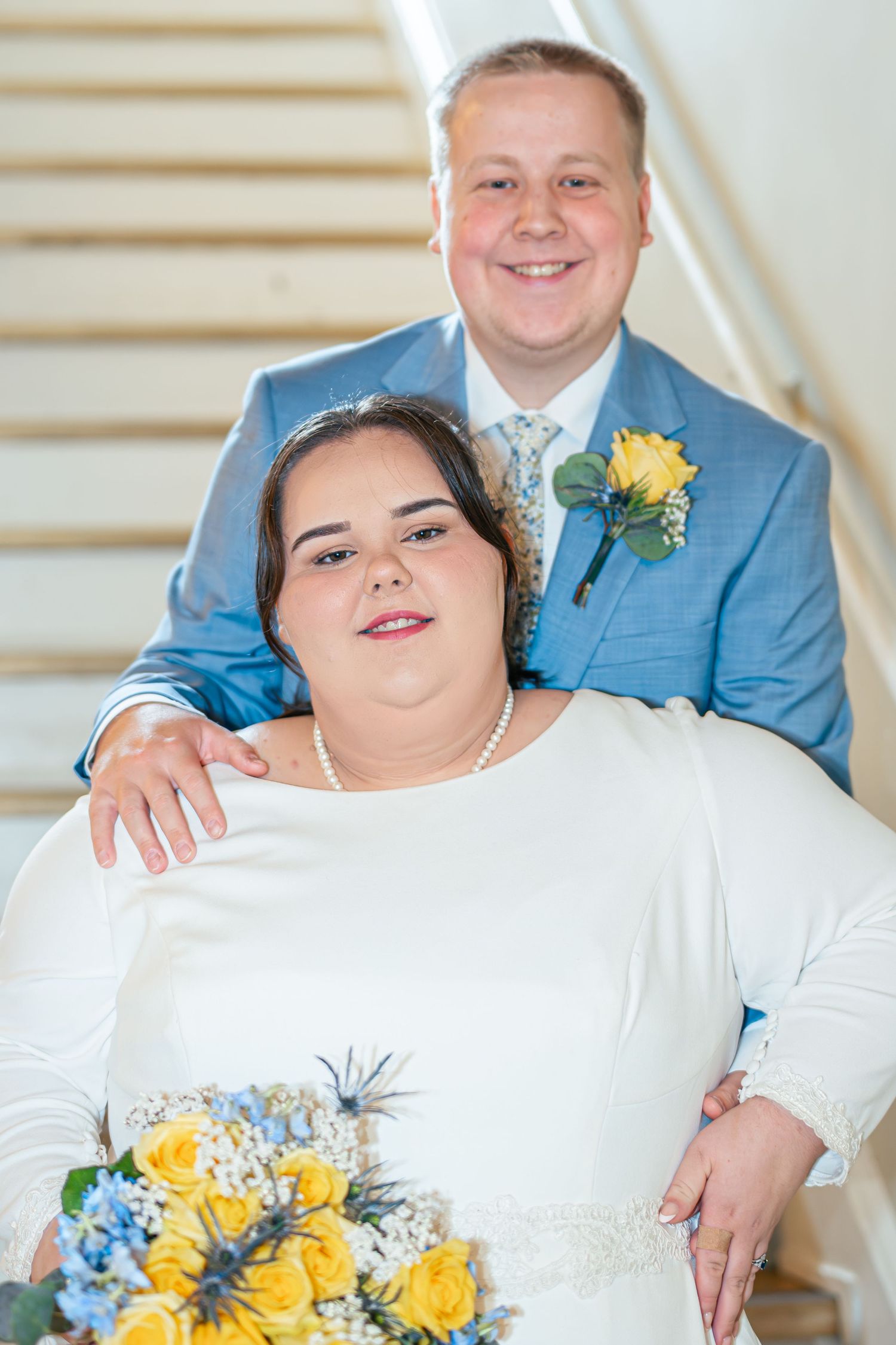 A wedding couple in blue and white attire poses together with a yellow and blue floral bouquet.