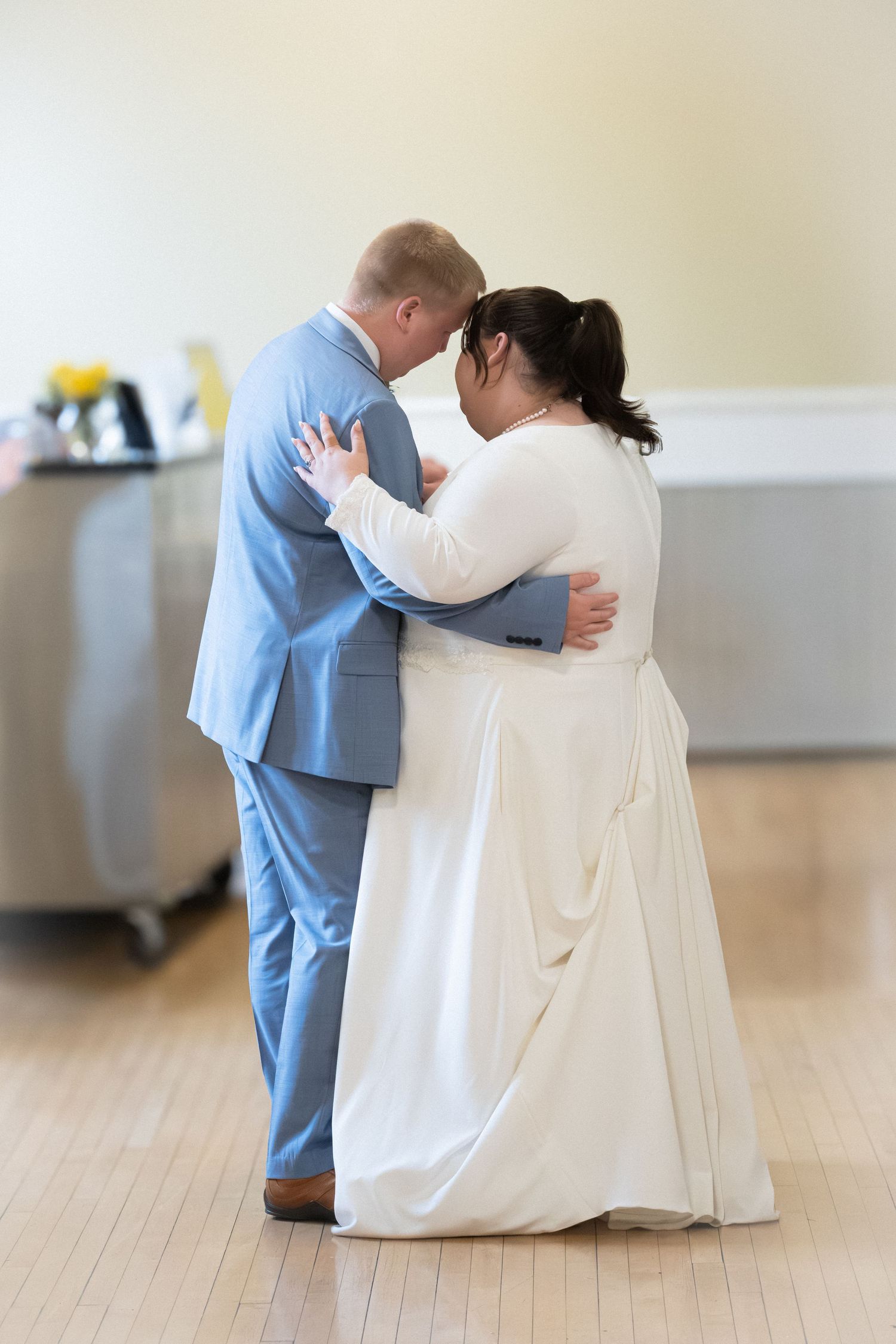 A couple in formal wedding attire share an intimate dance moment on a light wooden floor.