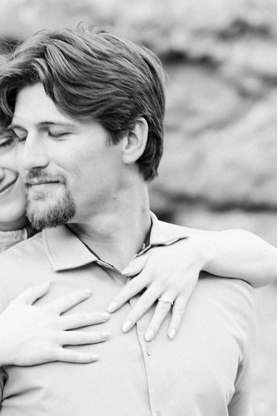 Black and white photo of a couple embracing, woman with ring wrapping arms around man from behind.
