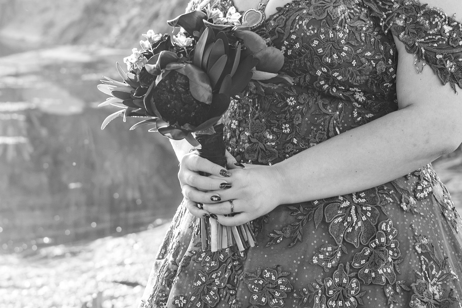 Black and white photo of a bride holding a floral bouquet, wearing an embroidered dress with a diamond ring visible.