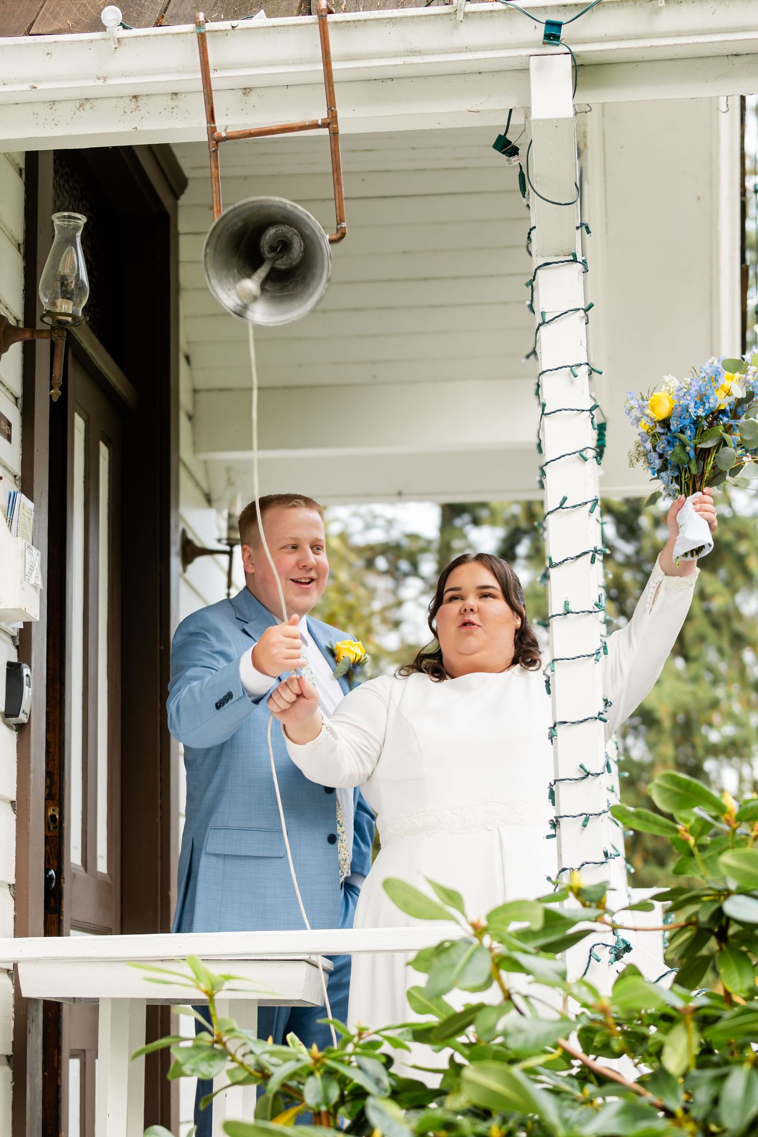 Two people laugh and celebrate together on a white porch decorated with string lights and greenery.