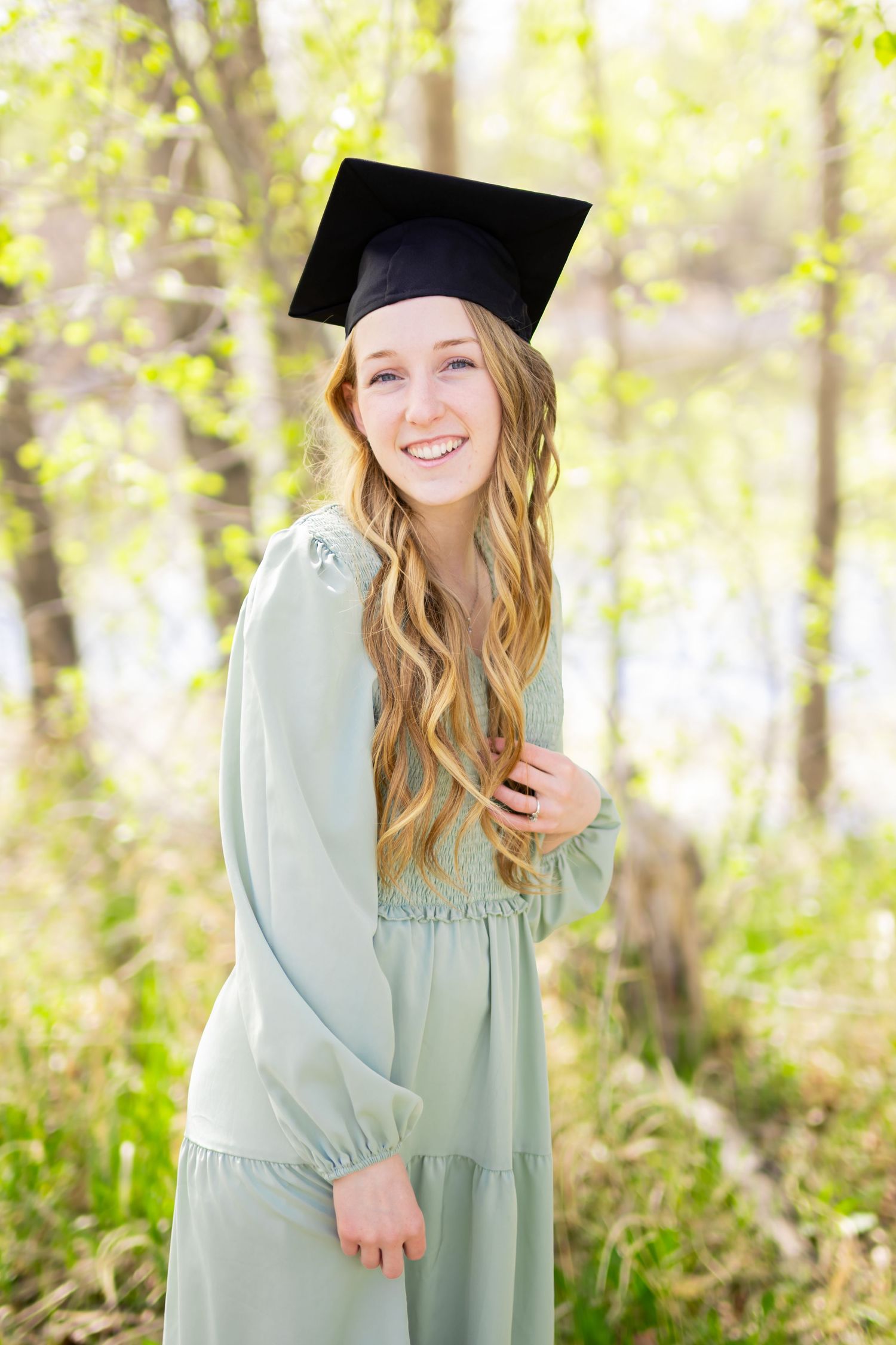 Smiling young woman in a black graduation cap and mint green dress posing outdoors among spring trees.