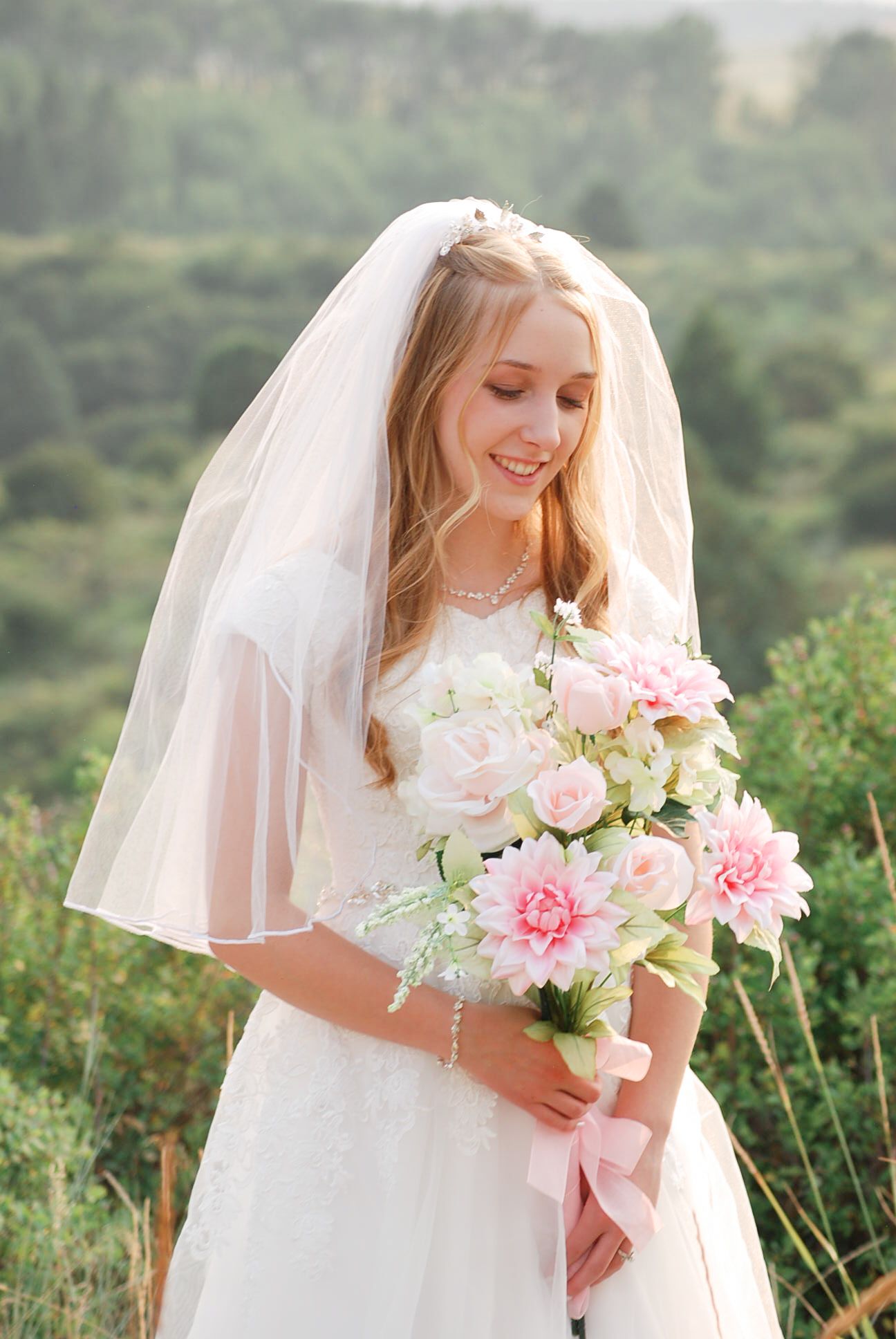 A bride in a white wedding dress and veil holds a beautiful pink and white bouquet in an outdoor garden setting.