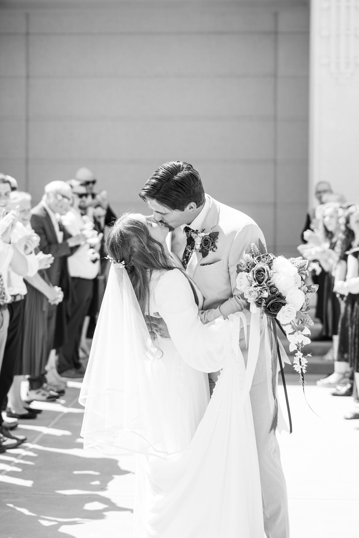Black and white photo of a bride and groom sharing their first kiss at the altar, surrounded by wedding guests.