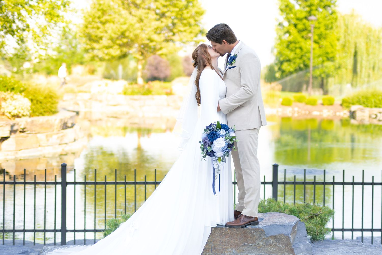 Bride and groom share a romantic kiss near a pond, bride holding blue bouquet, groom in tan suit, lush greenery backdrop.