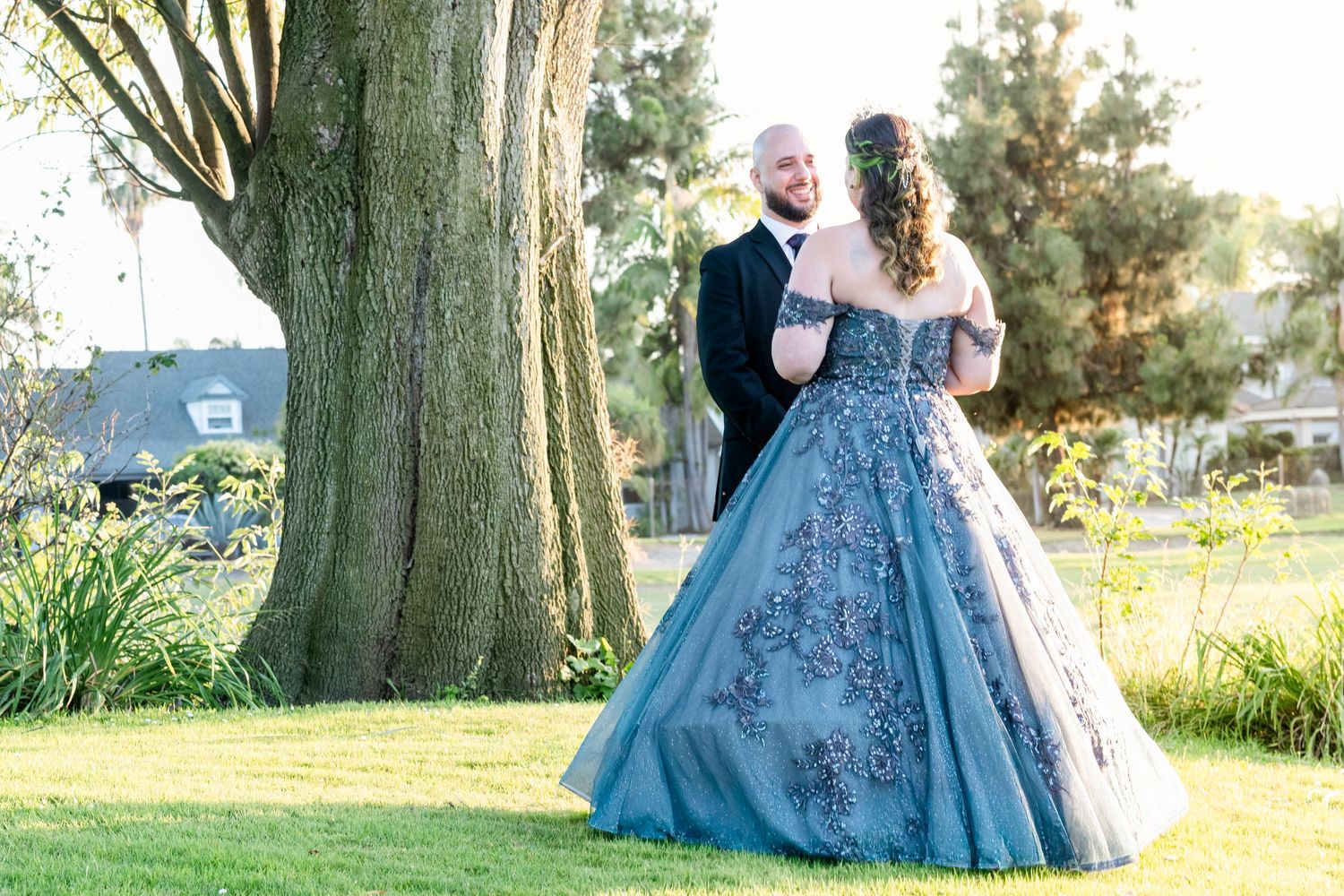 Couple in formal attire, woman in blue floral ball gown, man in black tux, posing near large tree outdoors.