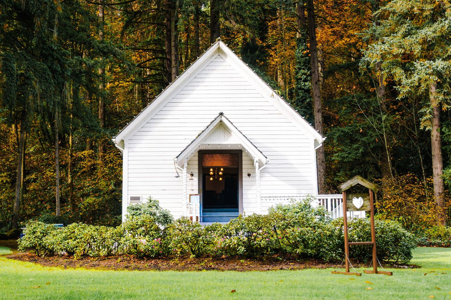 Small white chapel surrounded by lush green shrubs and autumn trees in a peaceful woodland setting.