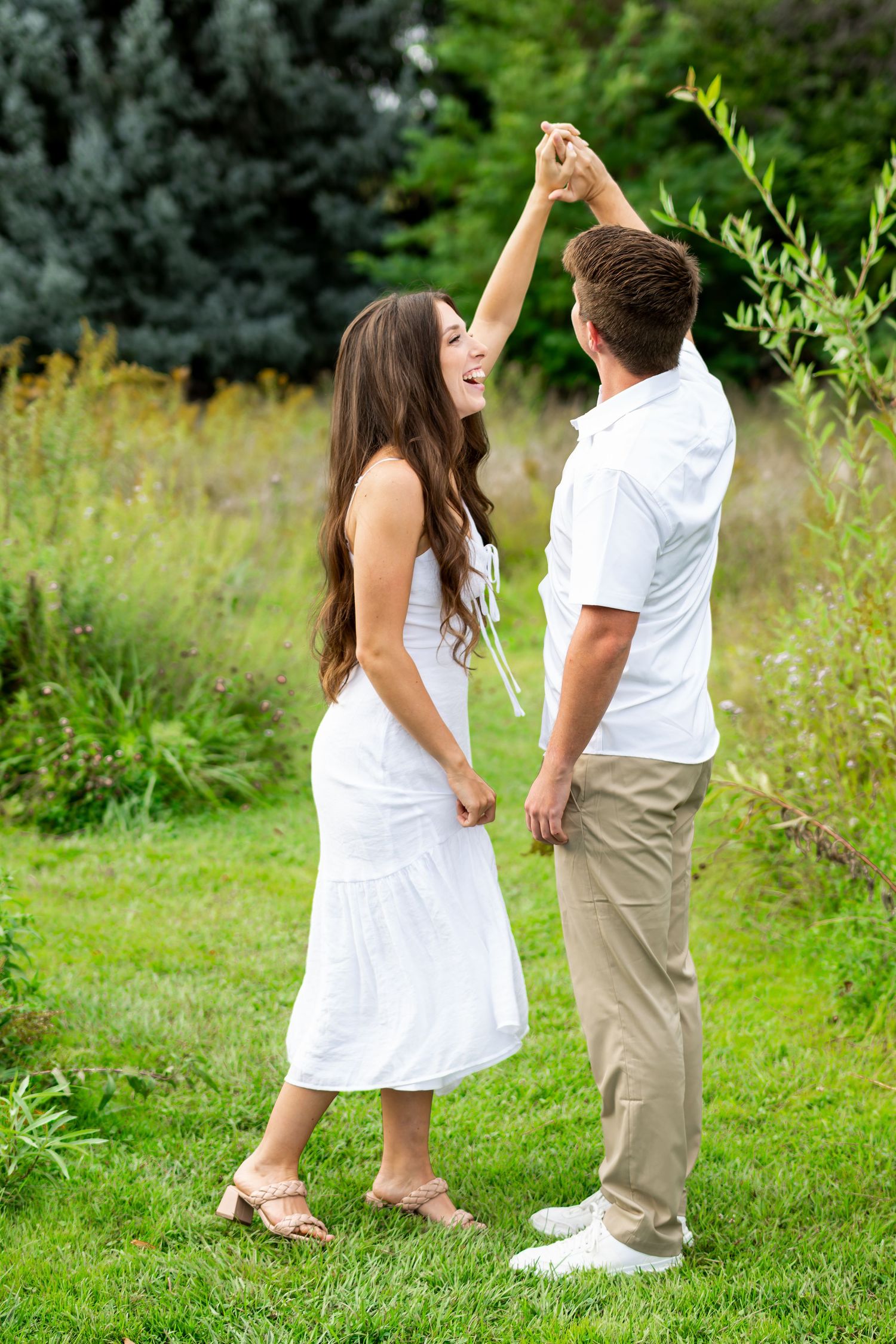 A couple dances together on green grass in a romantic outdoor setting, wearing white and khaki outfits.