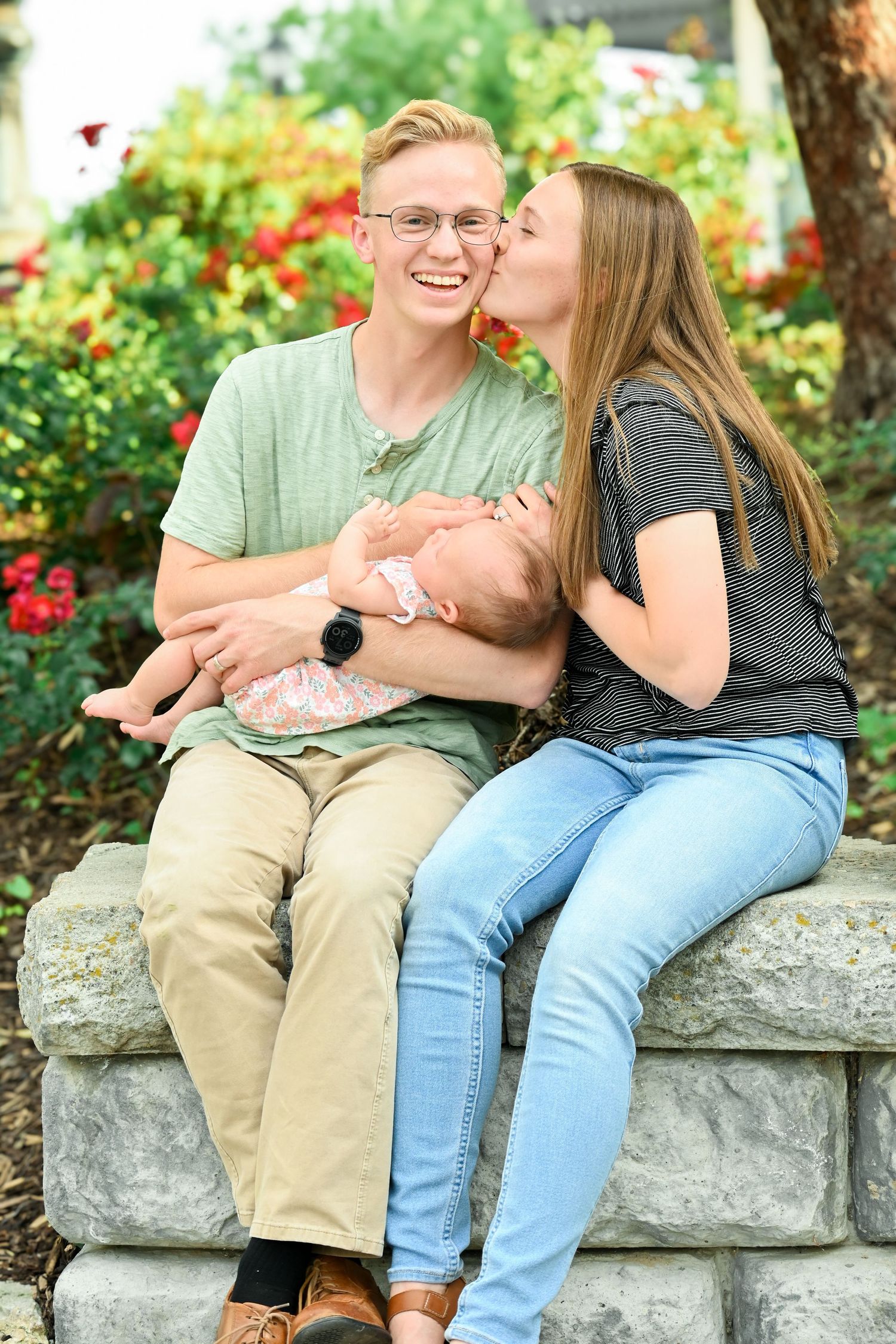 A couple with newborn baby sits together on a stone wall surrounded by blooming red flowers in a garden setting.
