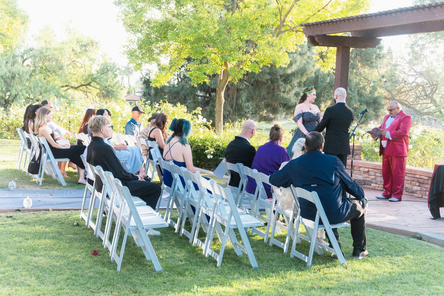 Outdoor wedding ceremony with guests seated in white folding chairs on green grass, surrounded by trees.