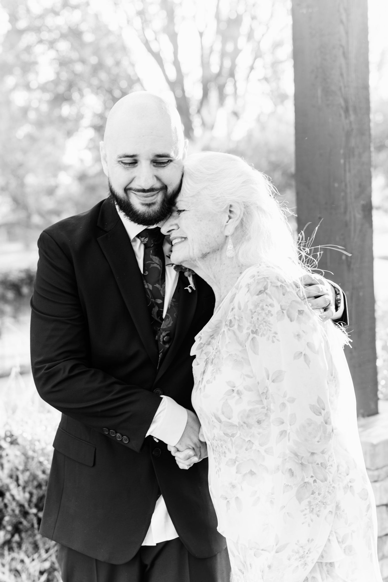 Black and white photo of a smiling bald groom in a suit embracing a bride in lace at an outdoor wedding.