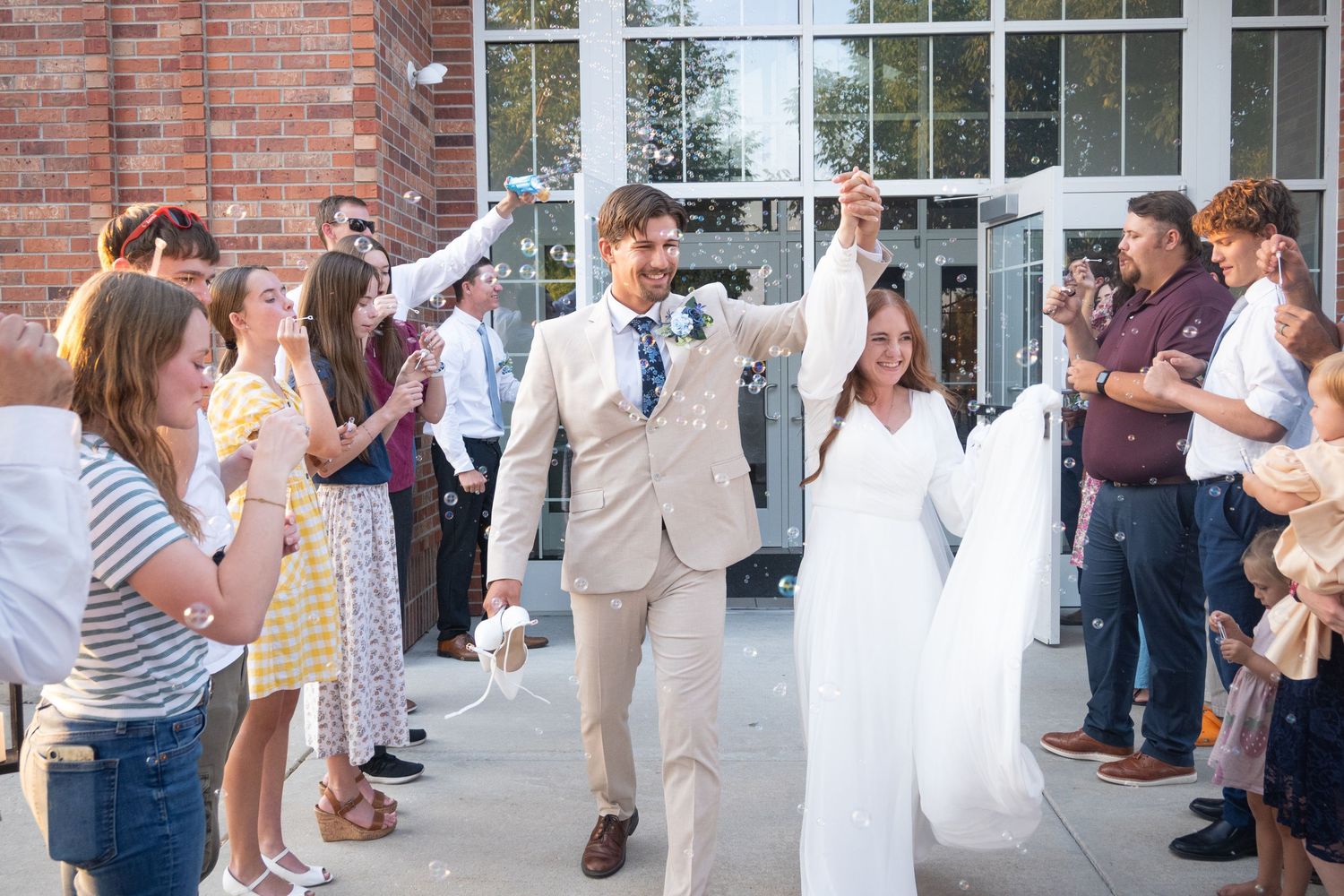 Bride and groom celebrate wedding exit with confetti, surrounded by cheering guests outside a brick building.