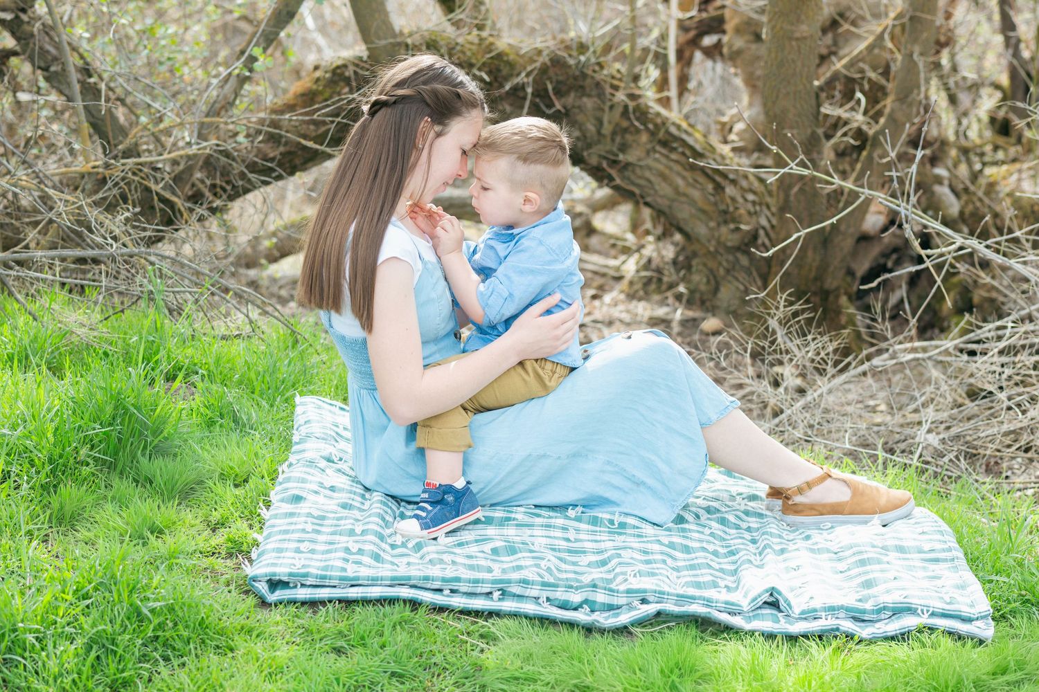 A mother sits on a blanket outdoors holding her toddler son, sharing a tender moment surrounded by green grass.