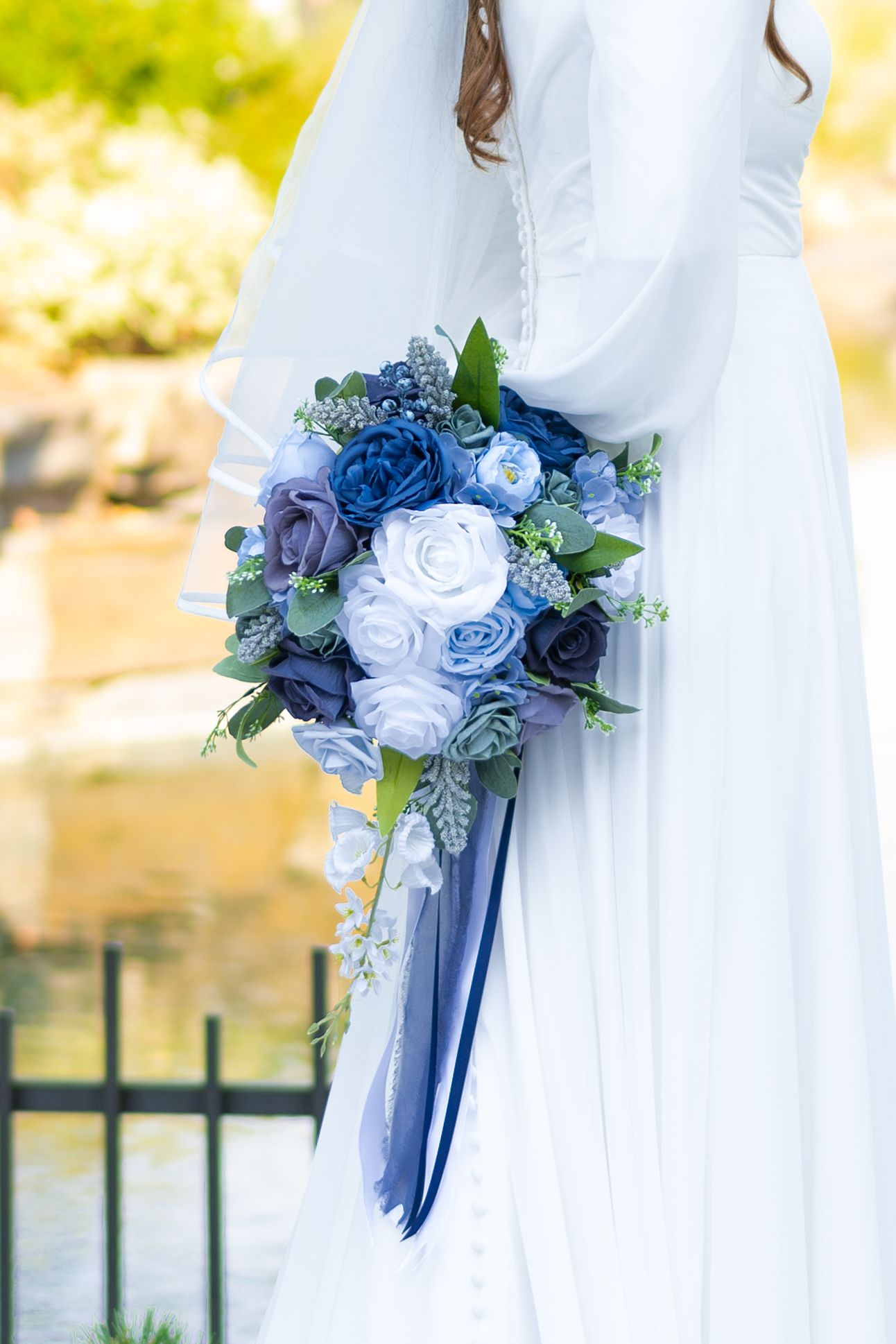 Bride in white gown holding cascading blue and white rose wedding bouquet with ribbon streamers outdoors.