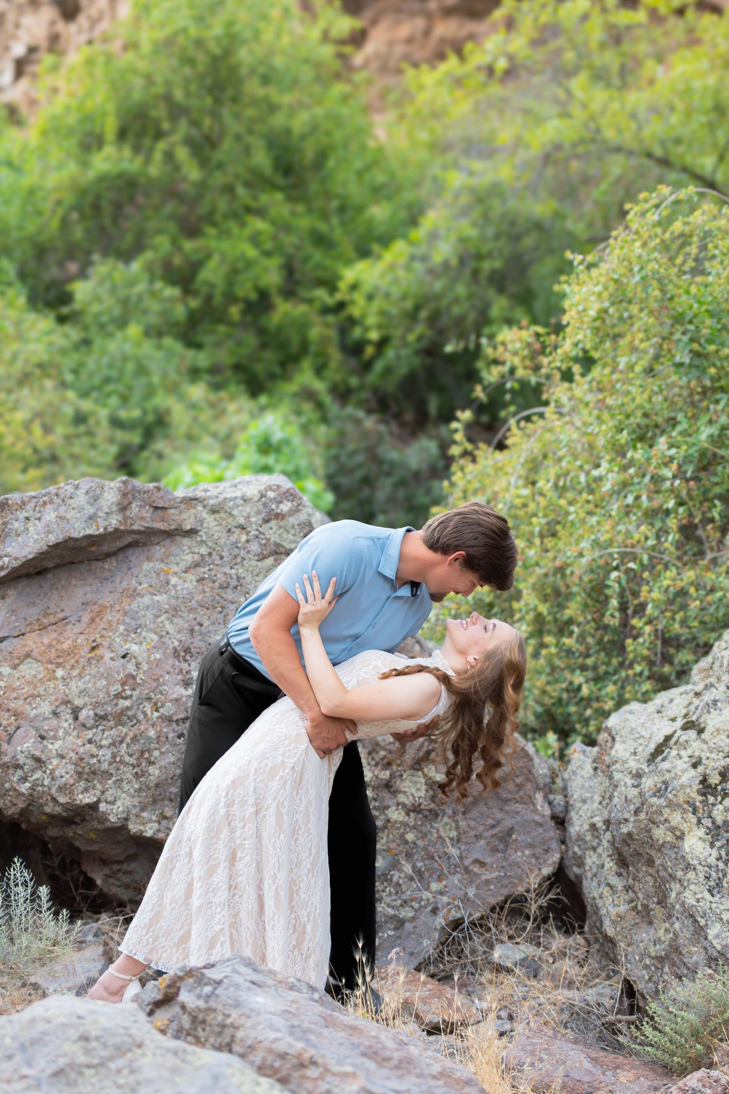 Couple sharing a romantic dip kiss among rocky boulders and lush green trees in an outdoor setting.