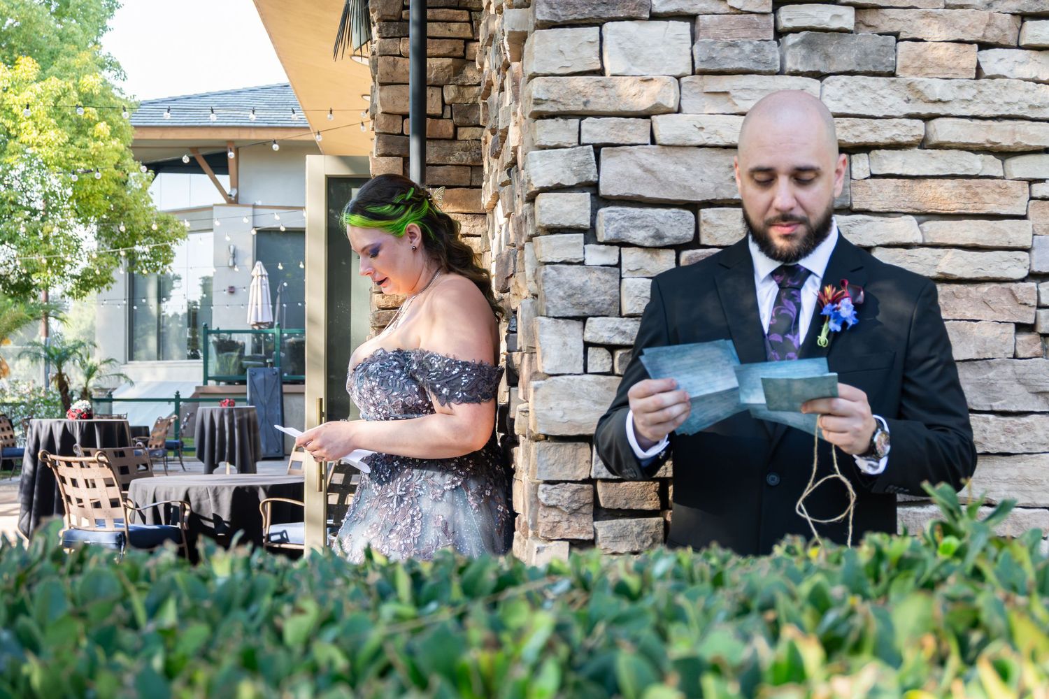 Bride and groom reading heartfelt letters before wedding ceremony outdoors near stone wall.