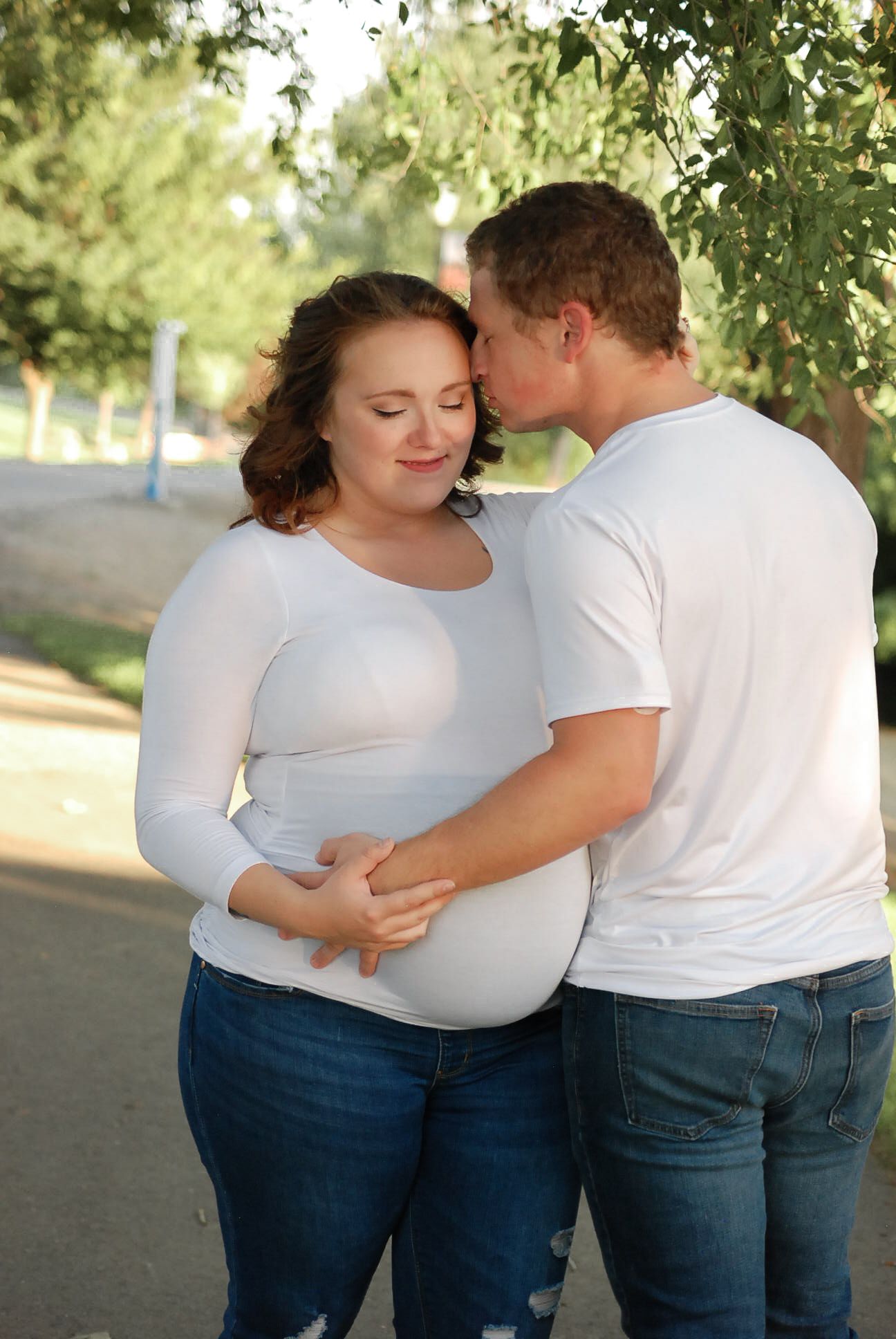 A couple in white shirts and jeans embraces lovingly under green trees.