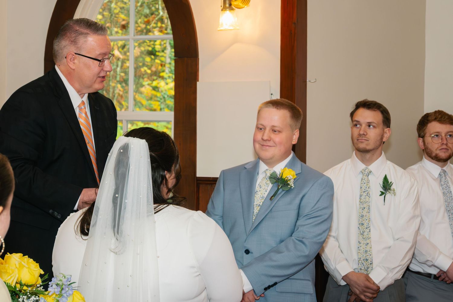 Groom in a blue suit with a yellow boutonniere smiles during a wedding ceremony as the bride in a veil and groomsmen look on.
