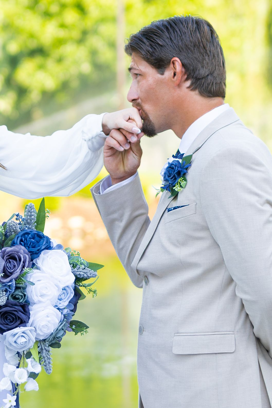 Groom in tan suit kissing bride's hand at wedding ceremony with blue and white floral bouquet.