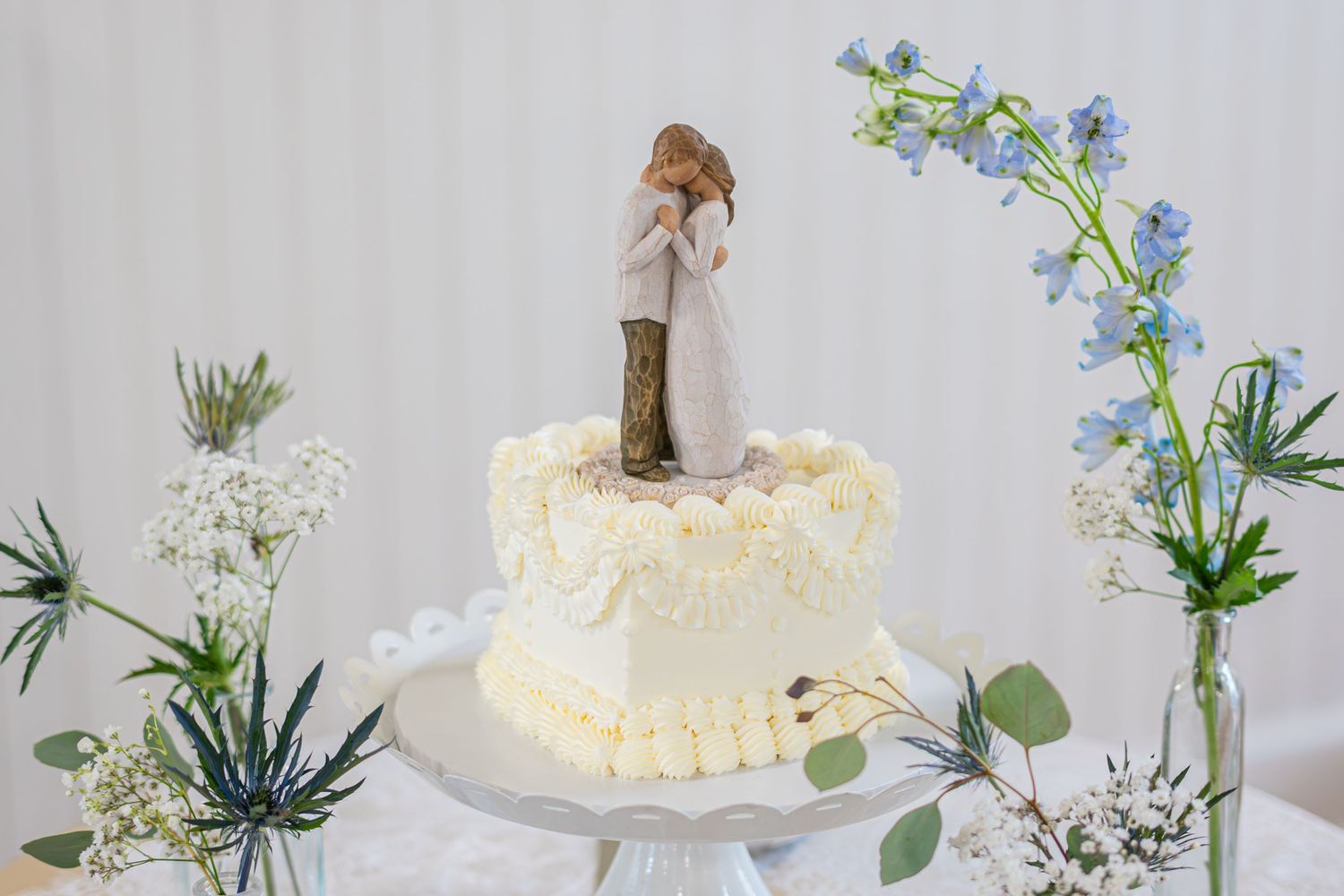 Elegant white wedding cake adorned with delicate blue flowers and greenery on a light gray background.