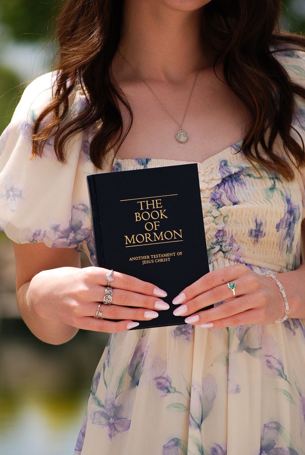 Person in floral dress holds The Book of Mormon while wearing multiple rings and a delicate necklace.