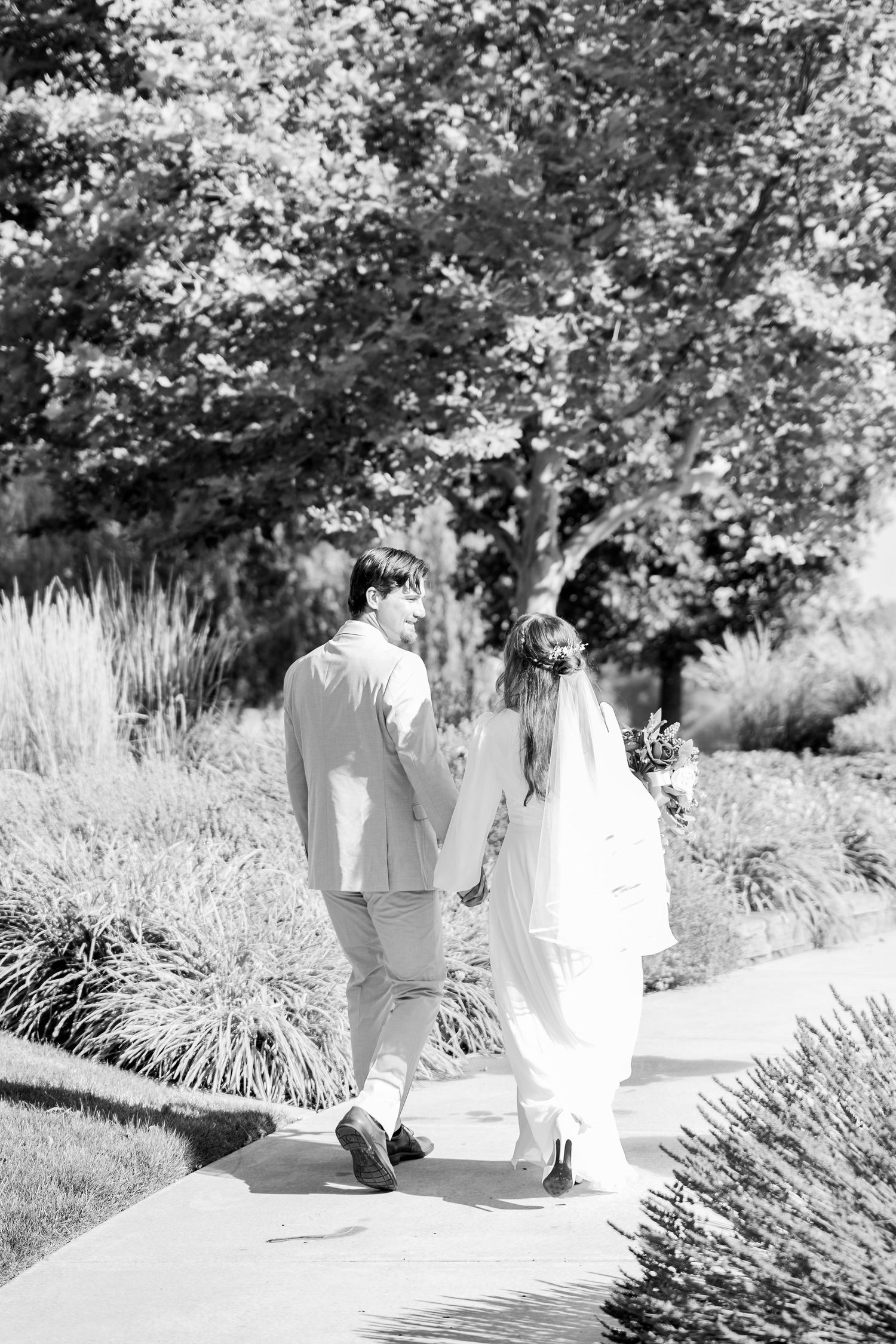 Black and white photo of bride and groom walking hand in hand along a garden path on their wedding day.
