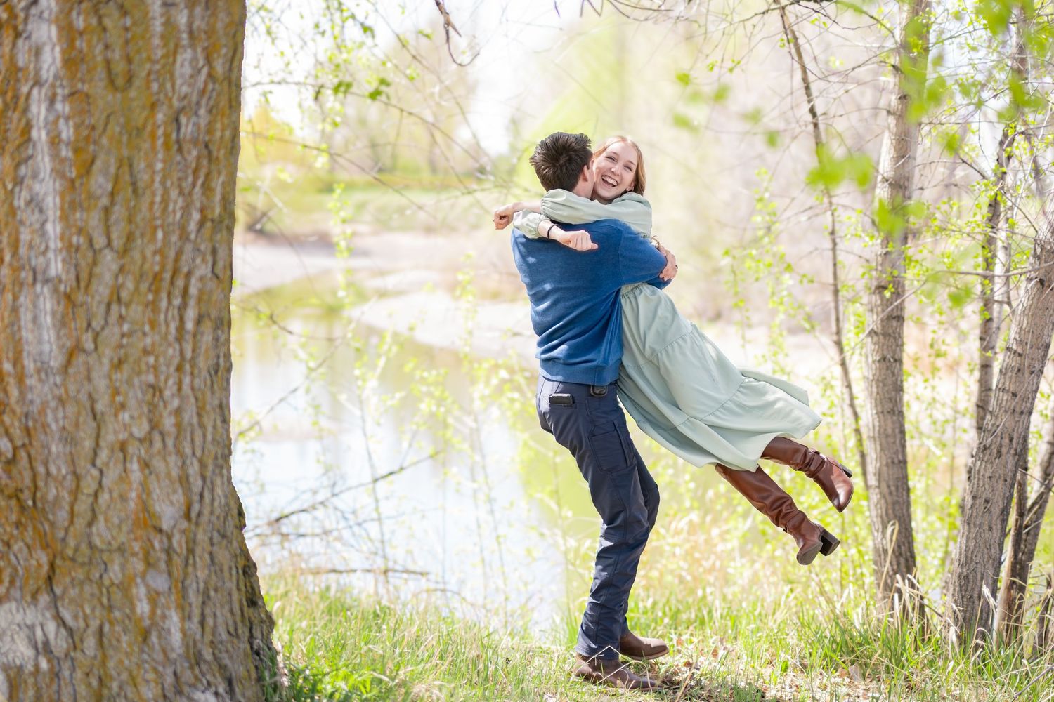 A man lifts and embraces a smiling woman outdoors near a serene pond surrounded by lush green trees.