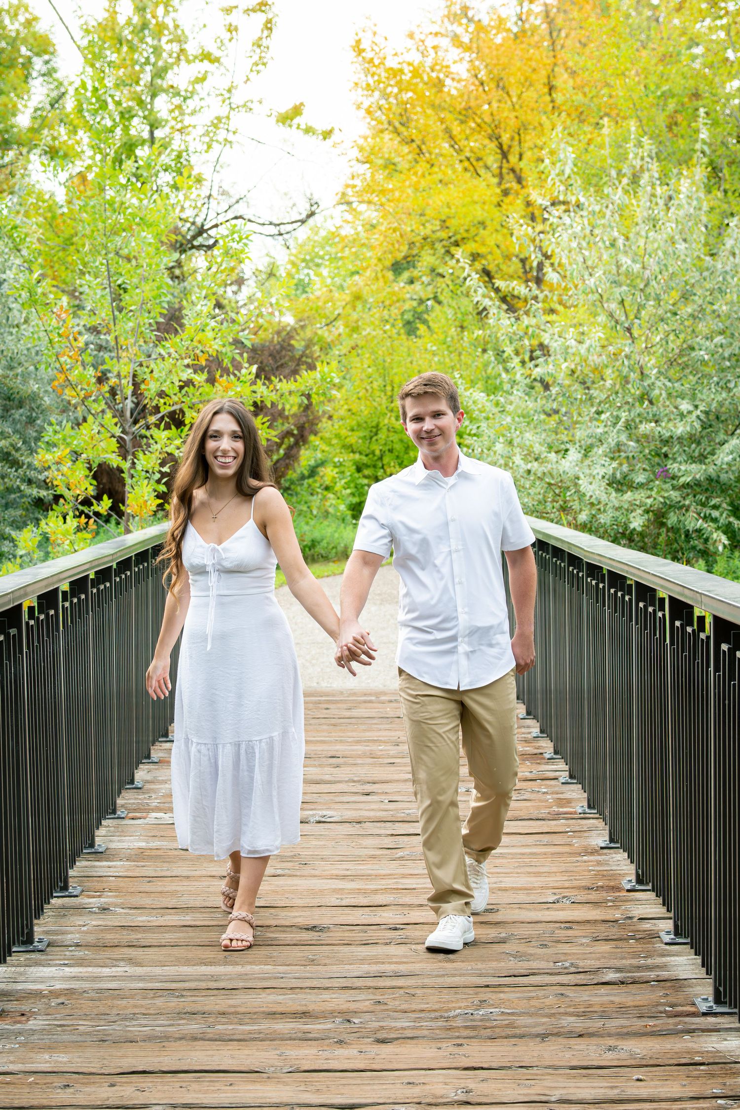 Couple in white and khaki walk playfully across wooden bridge surrounded by yellow trees.