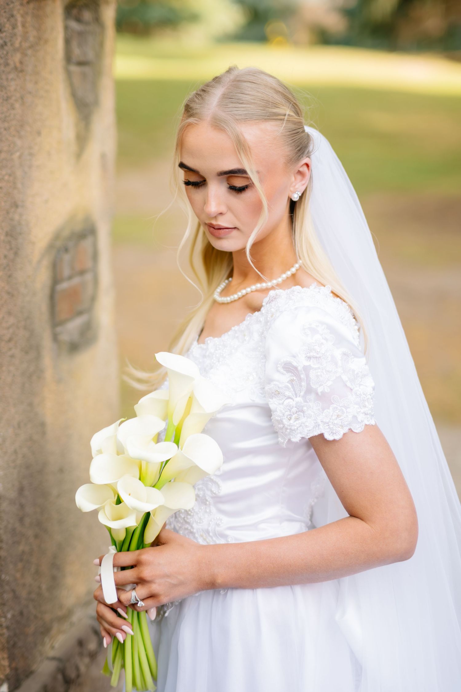 Blonde bride in white lace gown holding calla lily bouquet, wearing pearl necklace and veil outdoors.