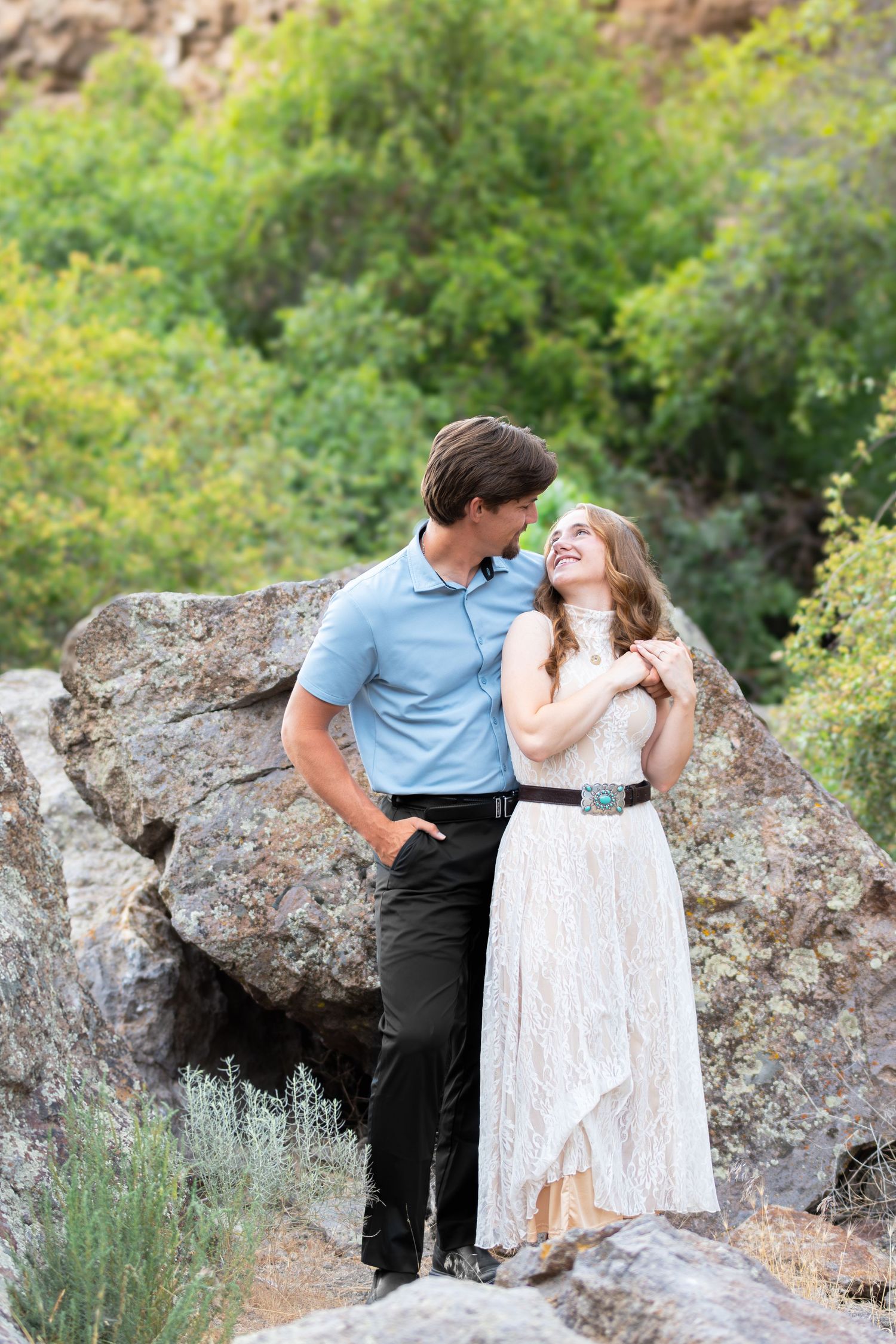 A smiling couple embraces on rocks outdoors, woman in white lace dress and man in blue polo shirt sharing a joyful moment.