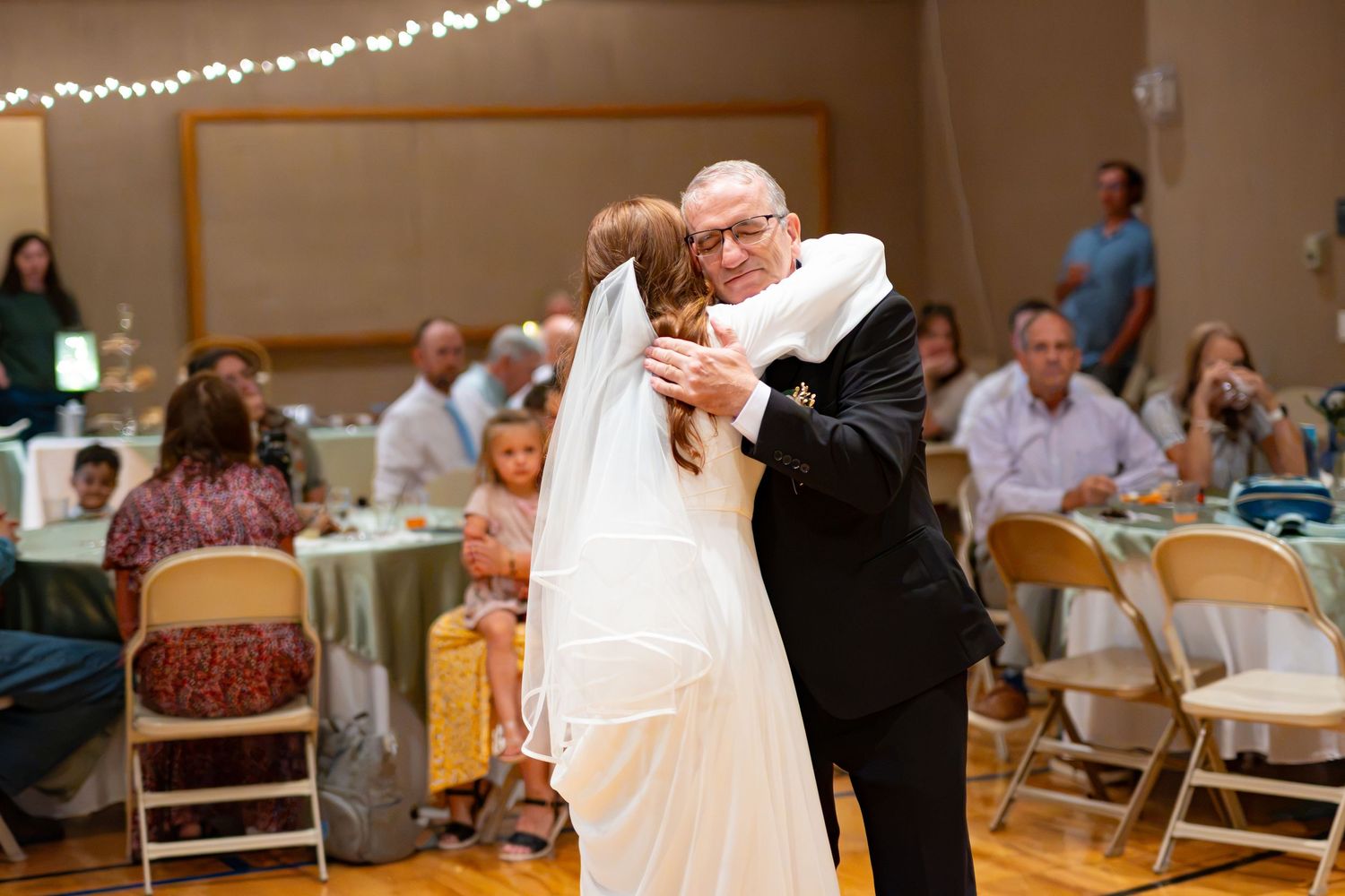 Bride and father share emotional first dance at wedding reception with guests seated at decorated tables in background.