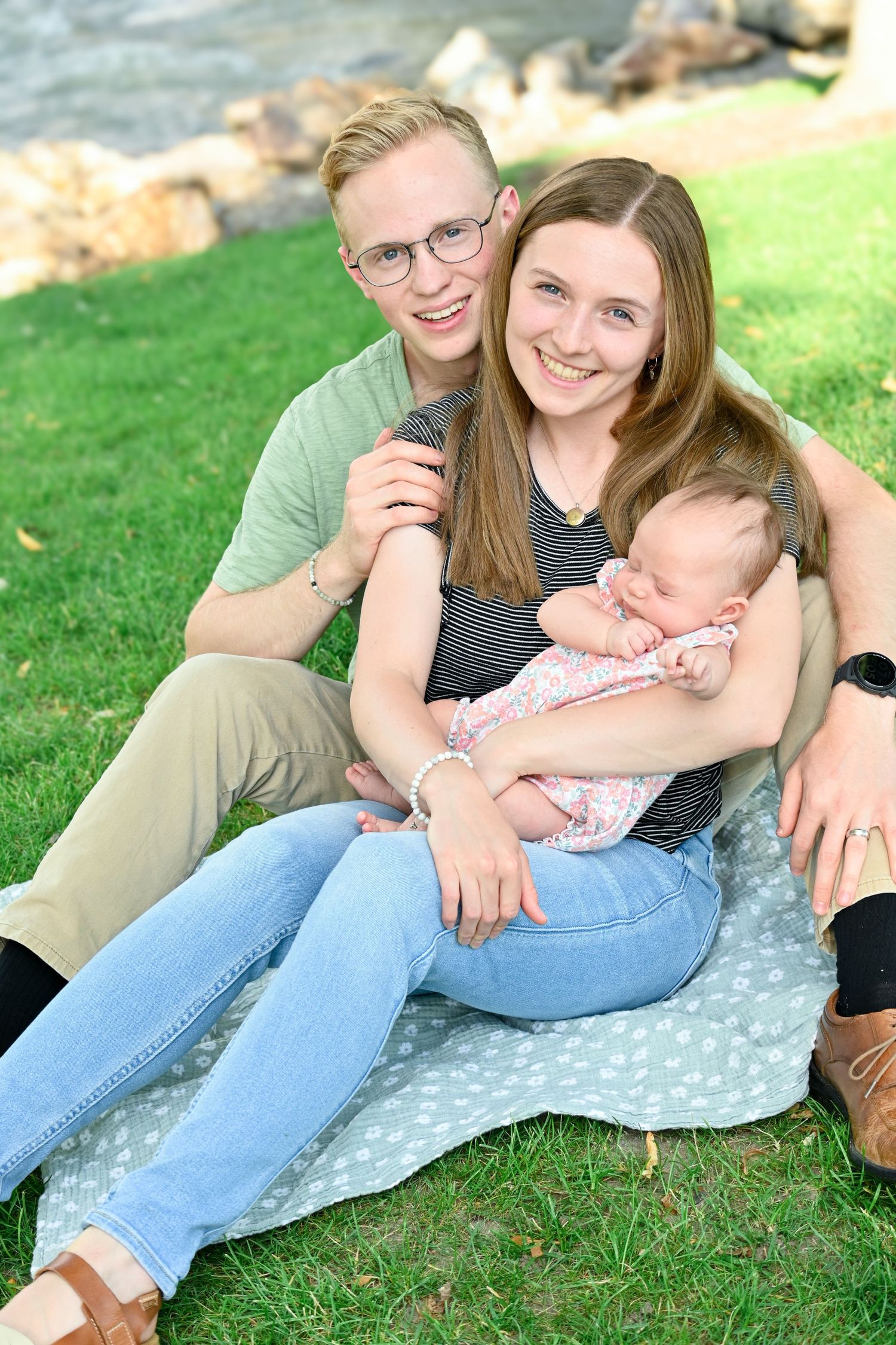 A family relaxes together on a blanket outdoors near rocky terrain and green grass on a sunny day.