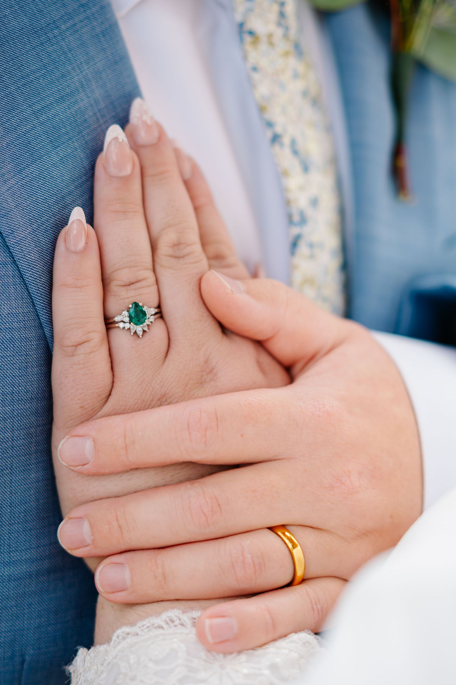 Close-up view of hands wearing wedding rings and an emerald engagement ring against blue and white fabric.