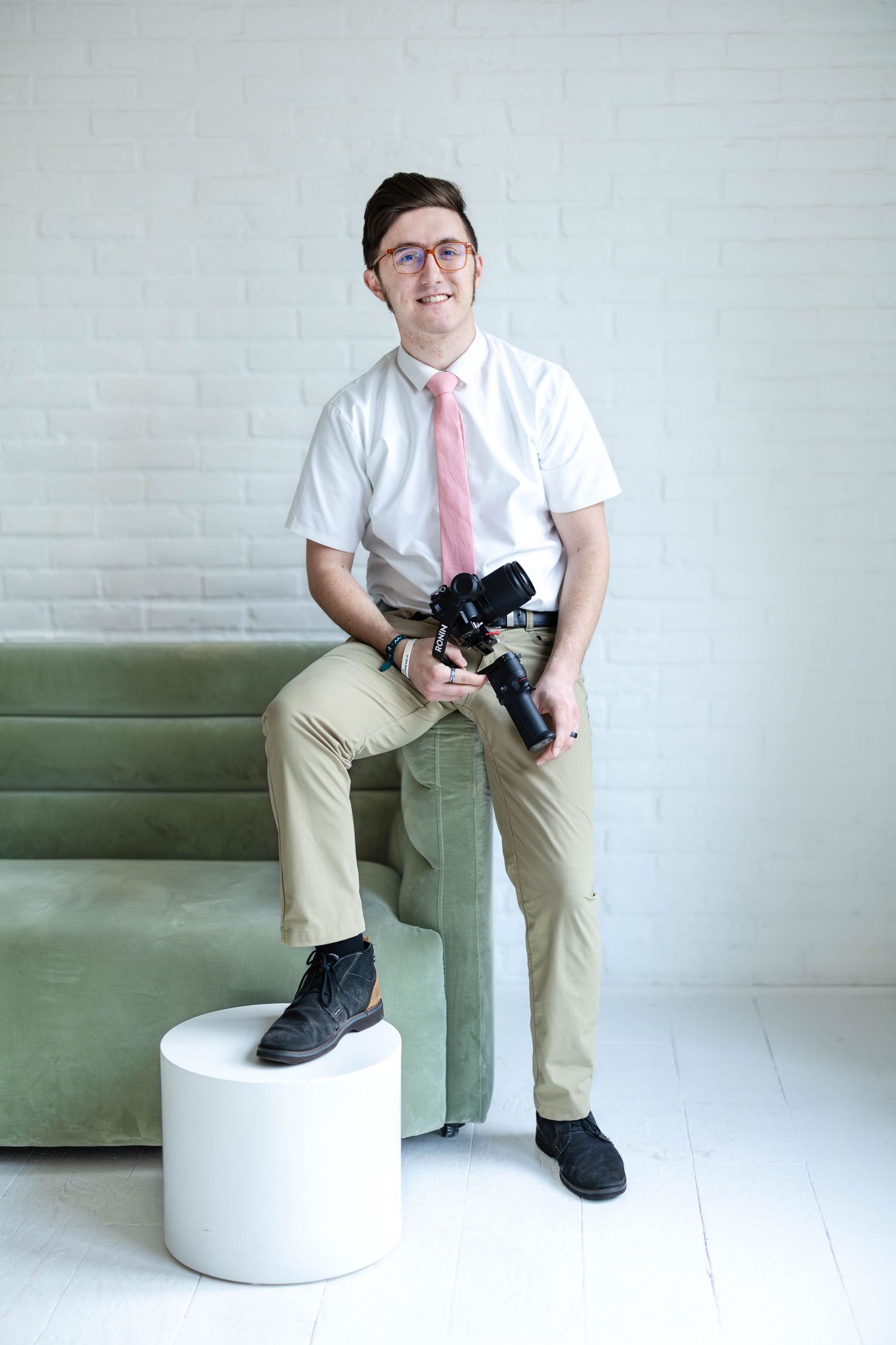 A person in business casual attire sits on a white ottoman while wearing black sneakers and a pink tie.