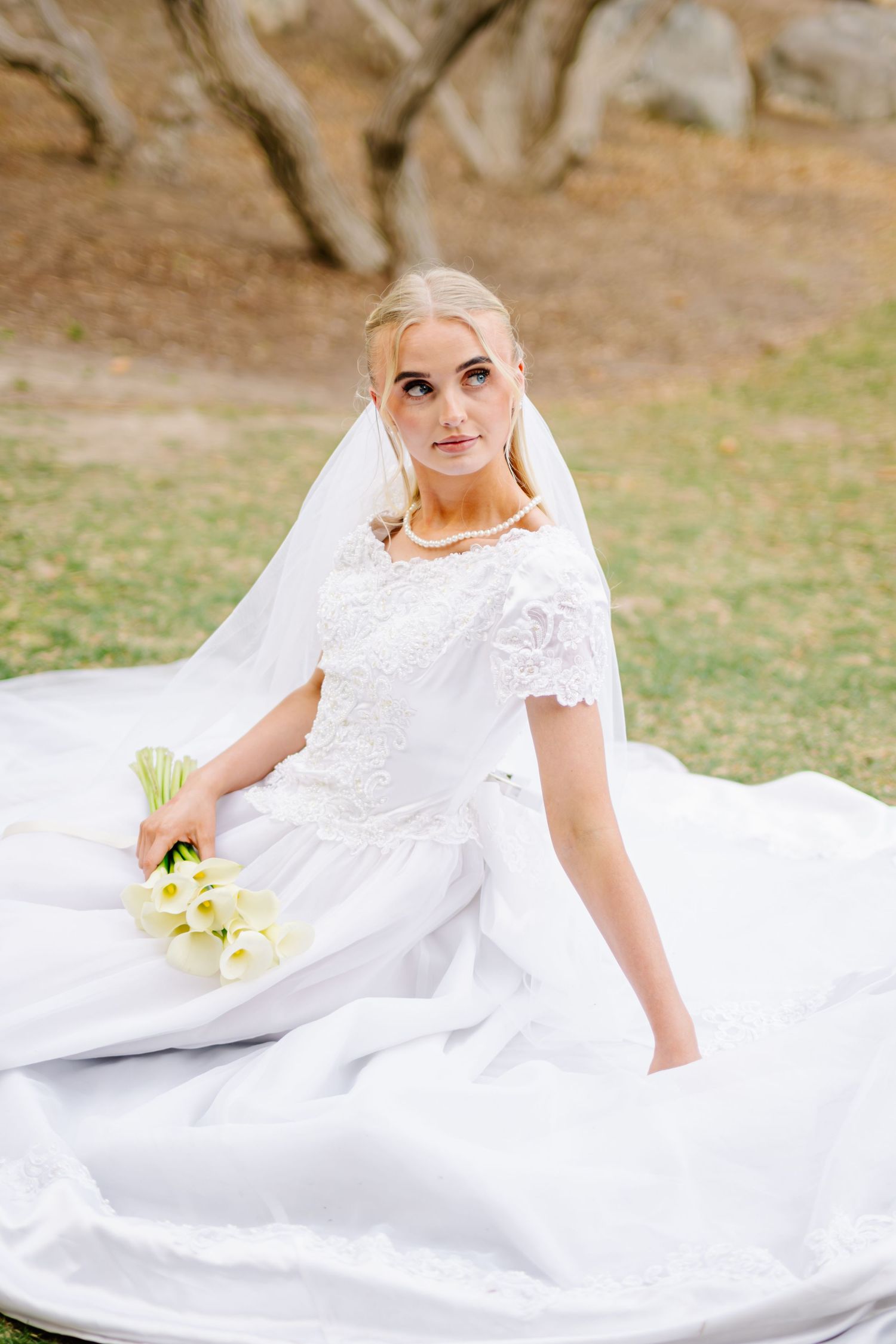 Elegant bride in white lace gown and veil sits outdoors holding calla lily bouquet on her wedding day.