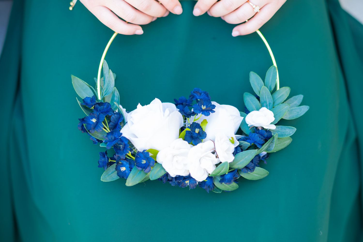 A person in a teal dress holds a gold hoop wreath adorned with white and blue flowers and green leaves.