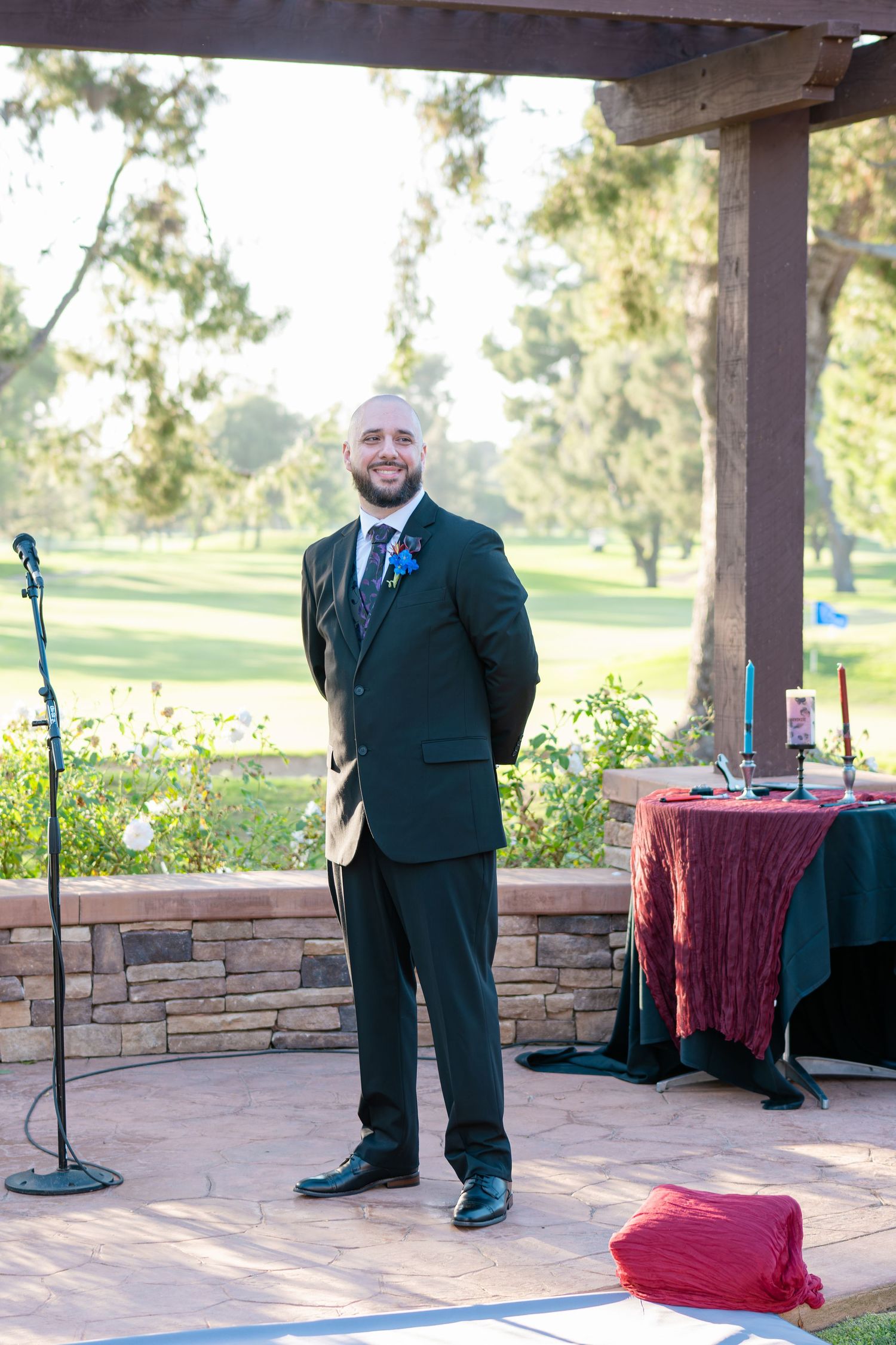 Smiling groom in dark suit with blue boutonniere stands at outdoor wedding reception venue with lush green golf course backdrop.