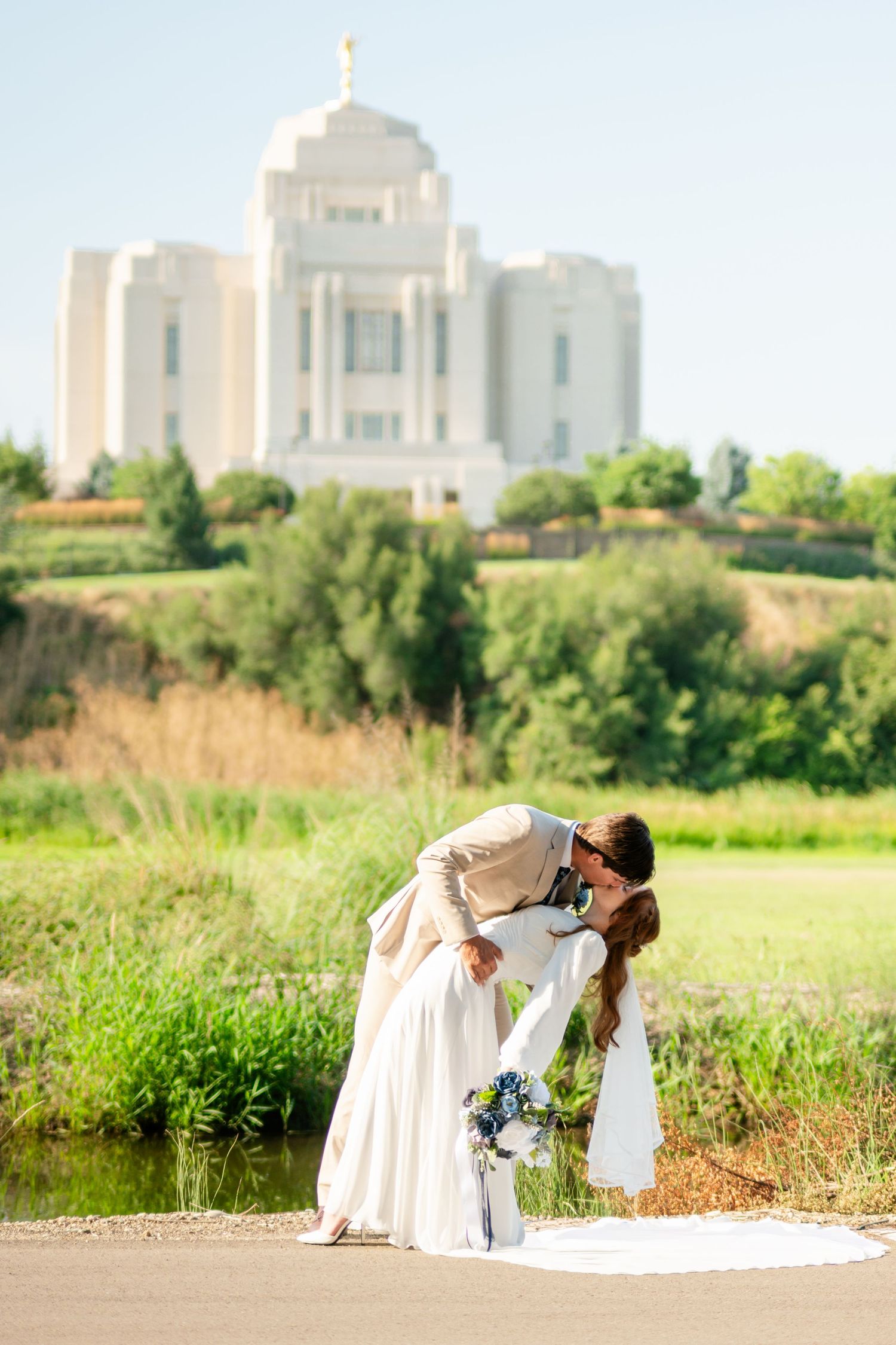 Bride and groom share a romantic dip kiss on a path with a white LDS temple on a hill in the background.