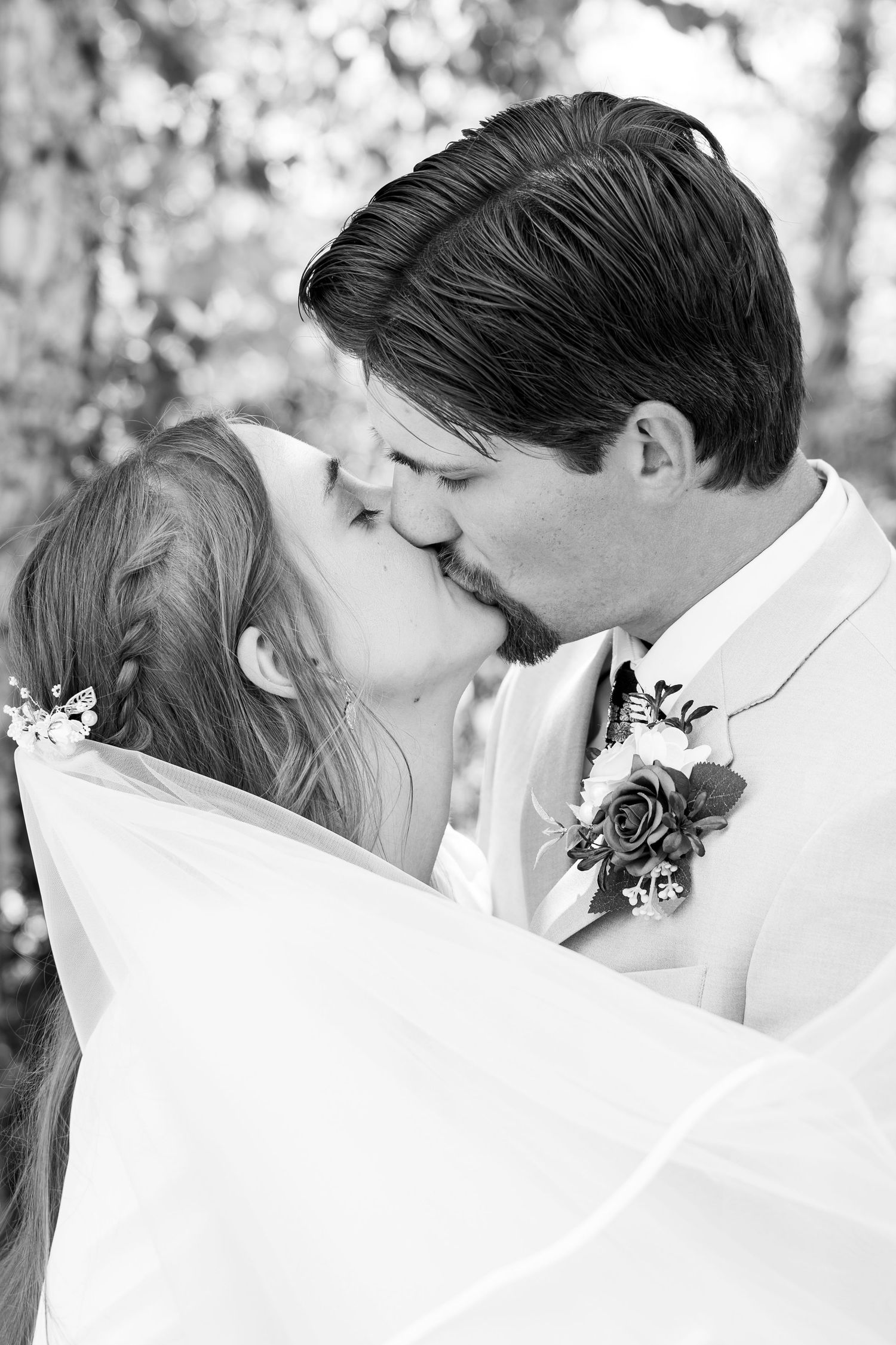 Black and white photo of a bride and groom sharing a romantic kiss, with veil flowing and groom wearing a floral boutonniere.