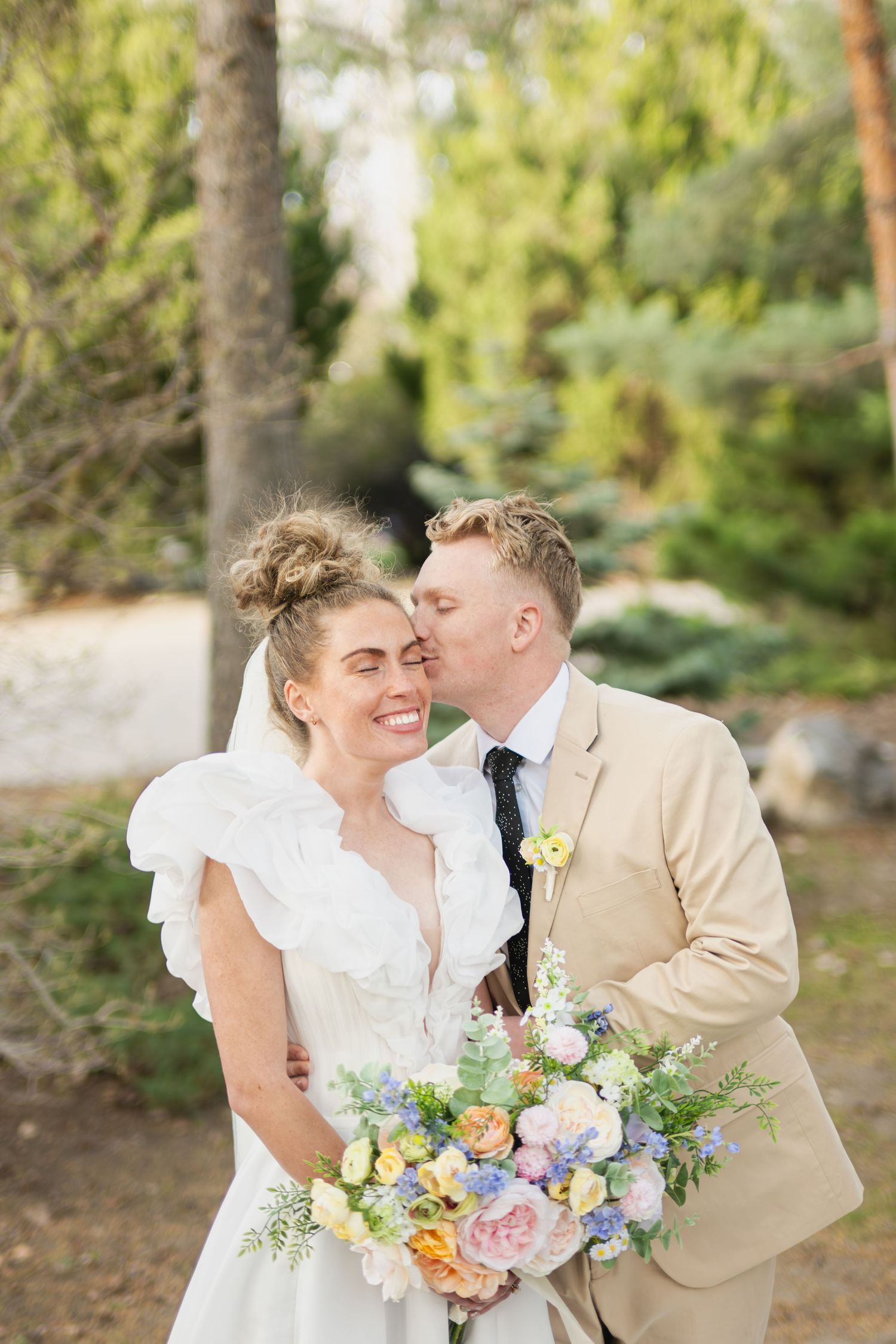 A romantic wedding couple shares tender moments in a sunlit forest setting with pastel floral bouquets.